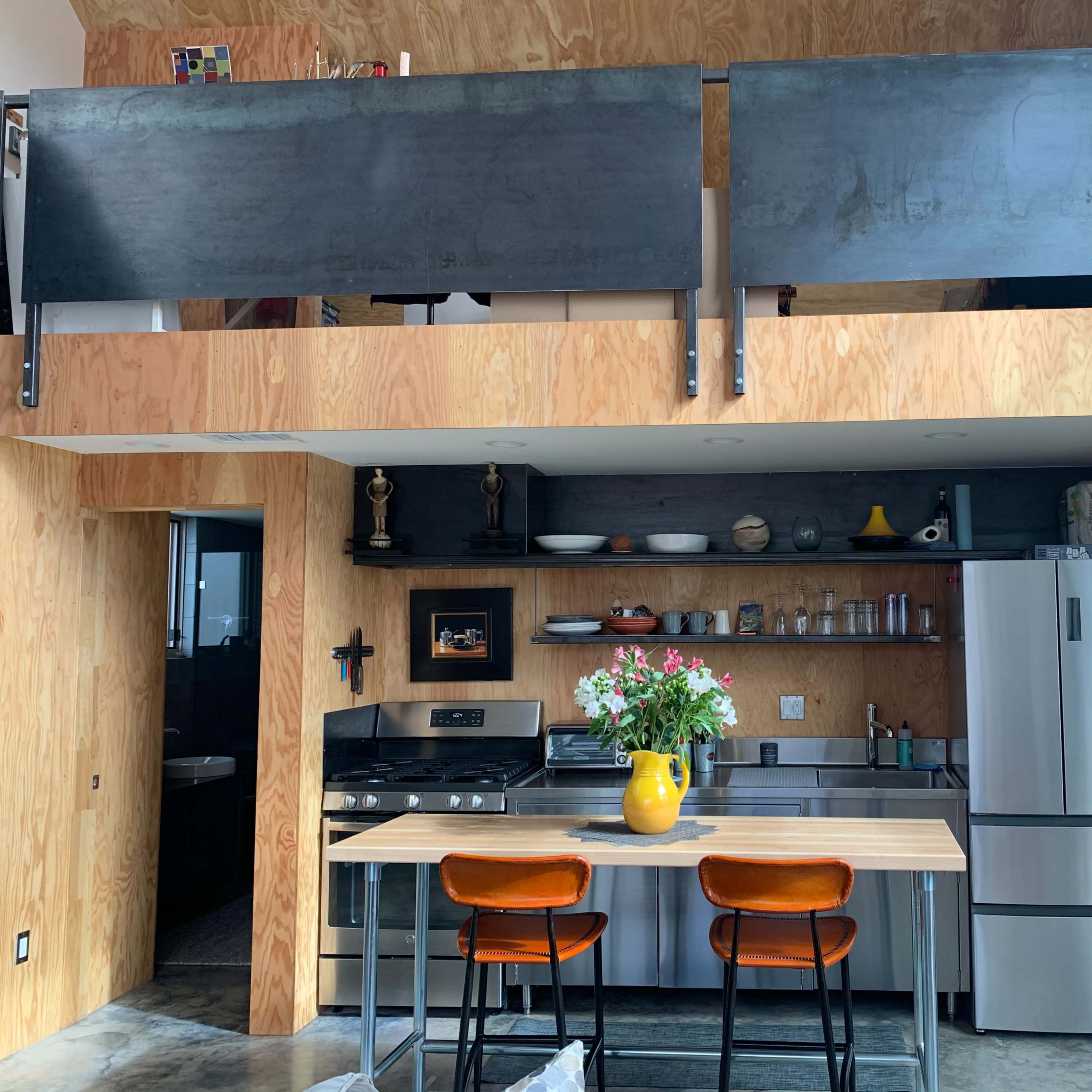 A modern kitchen with a central island, orange bar stools, and a balcony above, featuring wooden walls and stainless steel appliances.