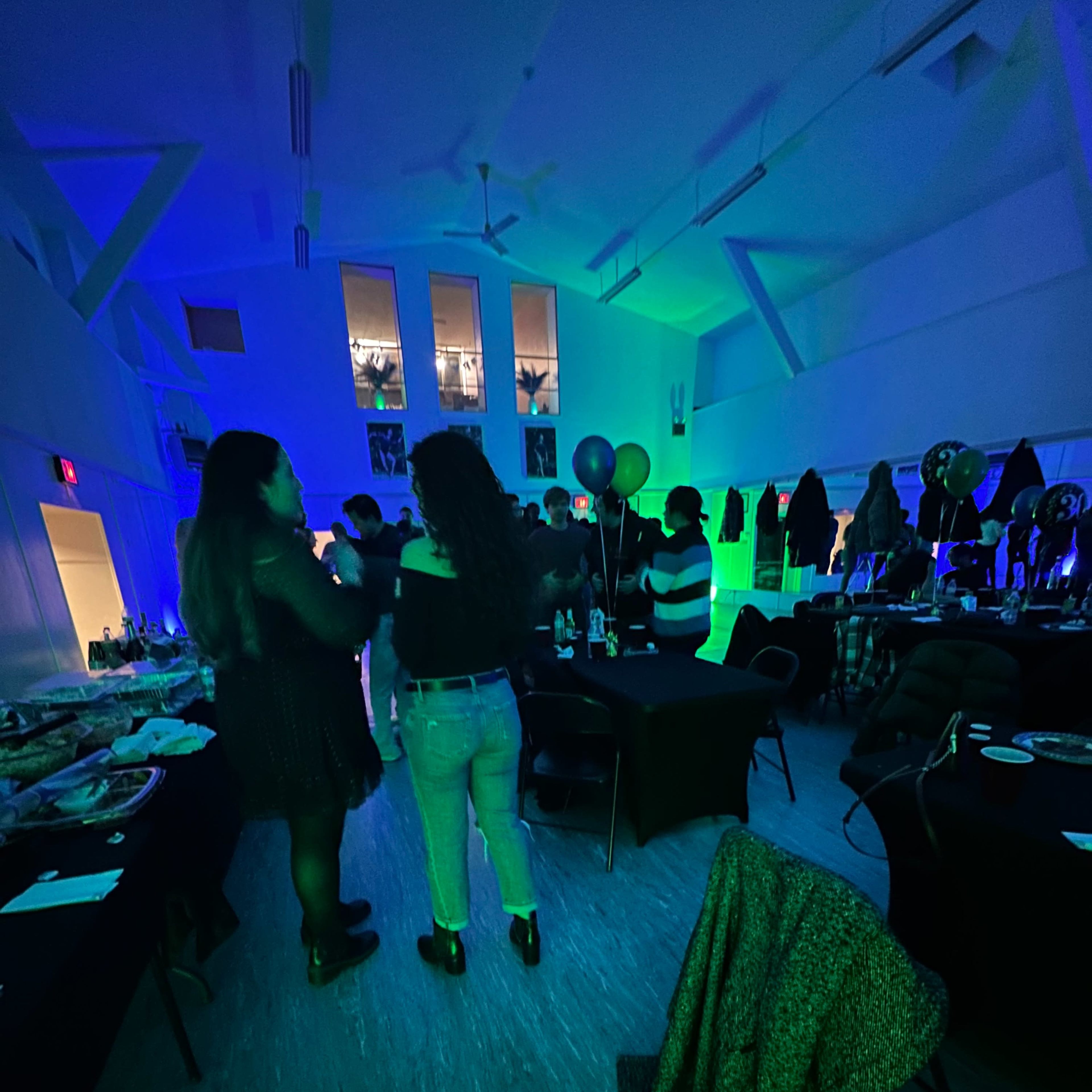 Two people are standing in a brightly lit room decorated for a party, surrounded by tables with food and drinks.
