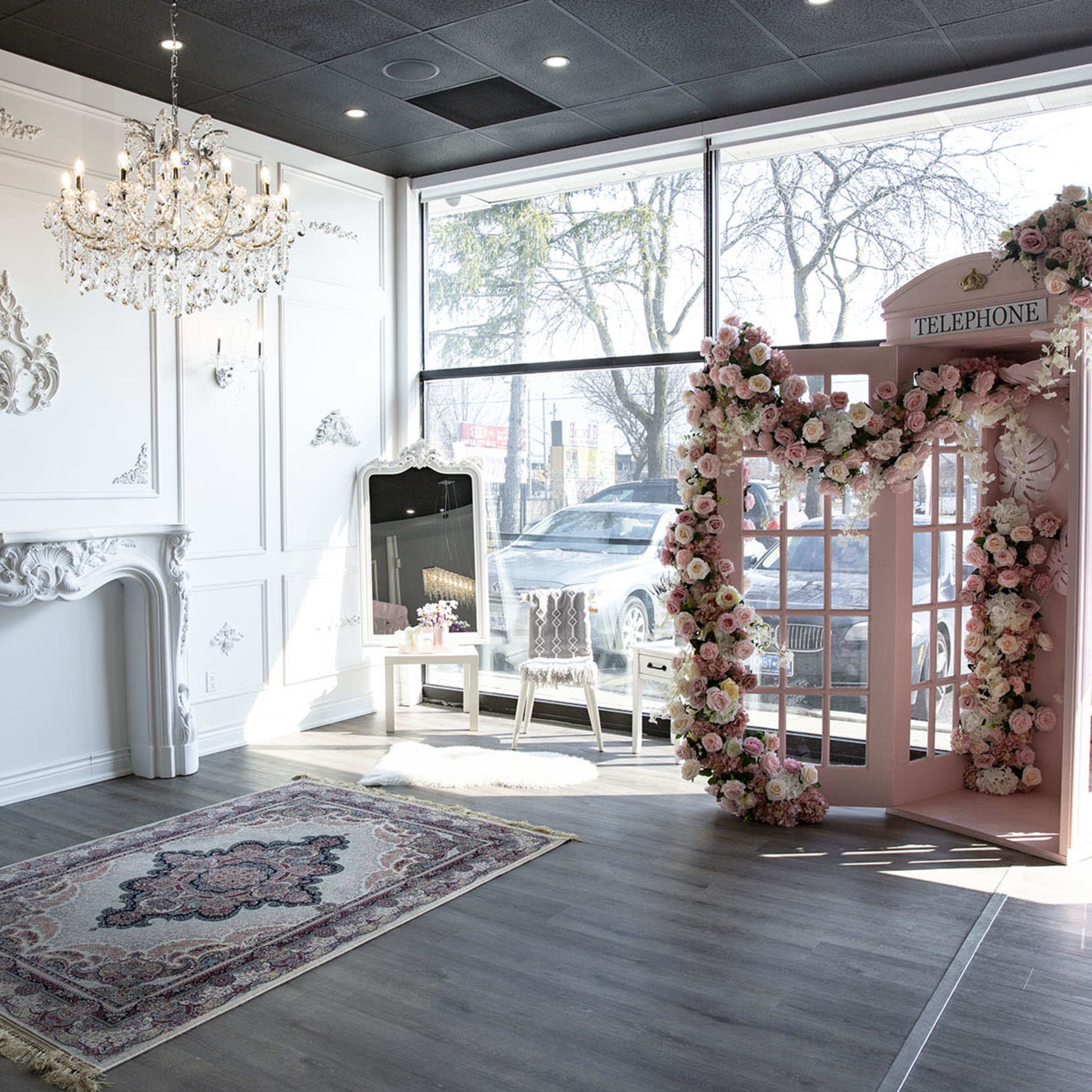 An elegantly designed event space featuring a pink floral archway resembling a vintage telephone booth, with a chandelier and large windows allowing natural light to enter.