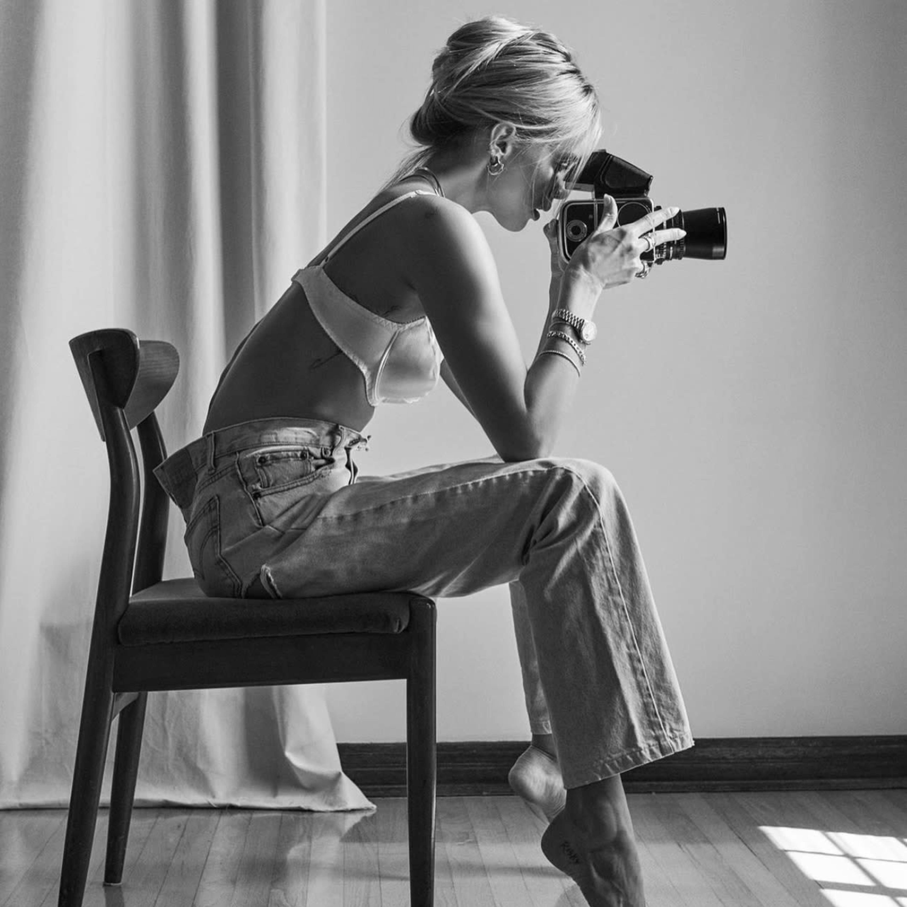 A woman sits on a chair, holding a vintage camera and focusing on a subject in a softly lit room.