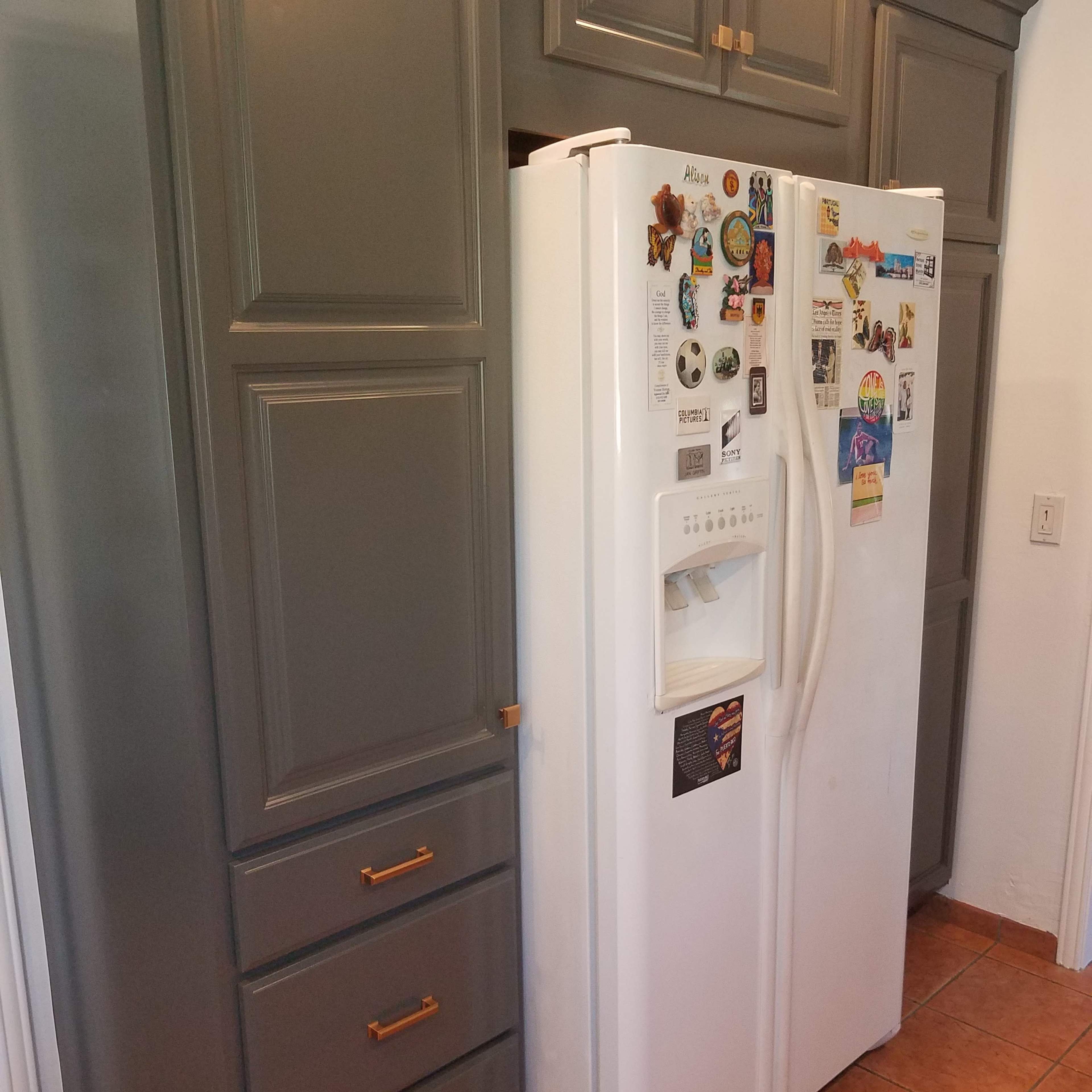A white refrigerator located between grey cabinets and drawers in a kitchen space with tiled flooring.