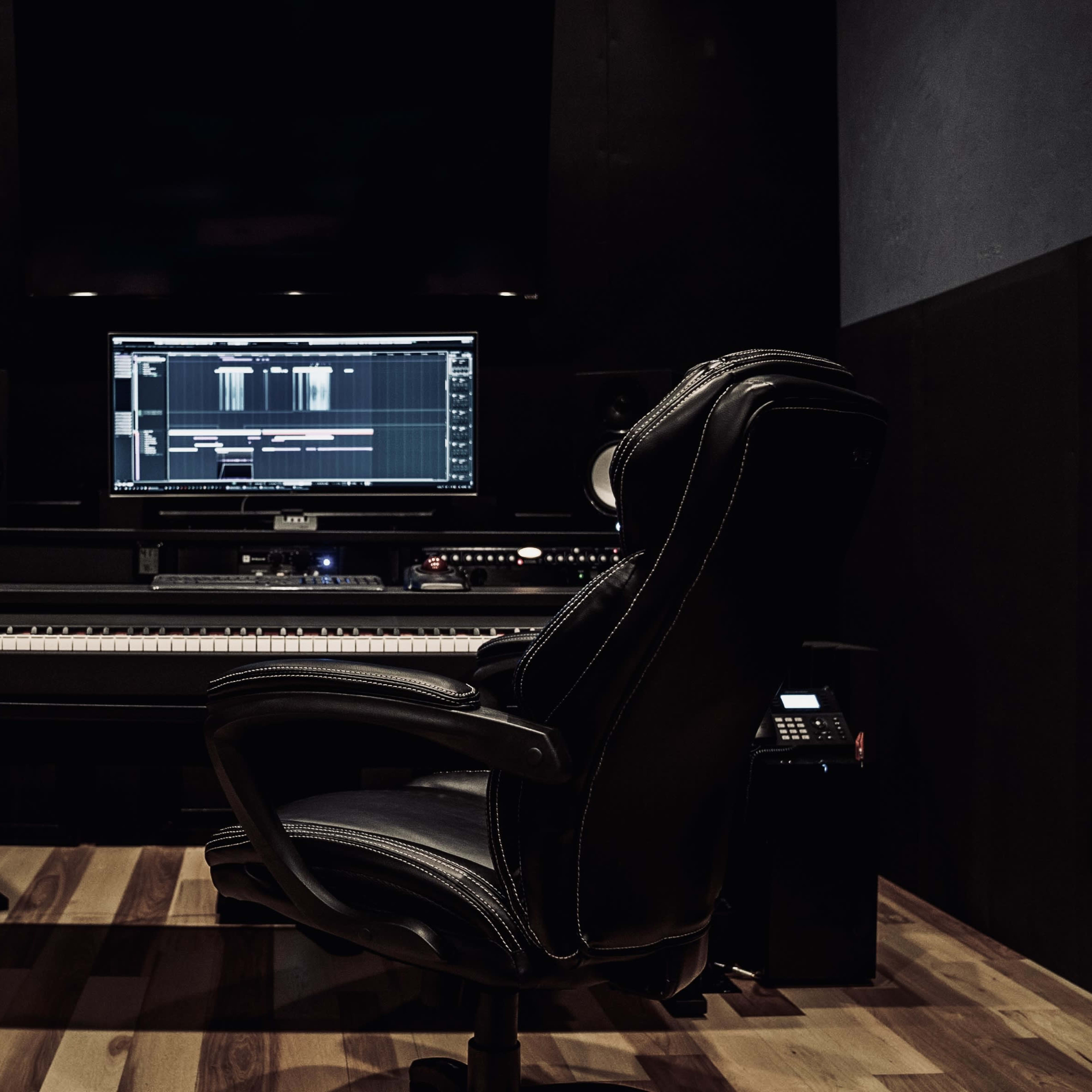 A professional music production studio features a digital audio workstation on the screen, a keyboard, and a black ergonomic chair.