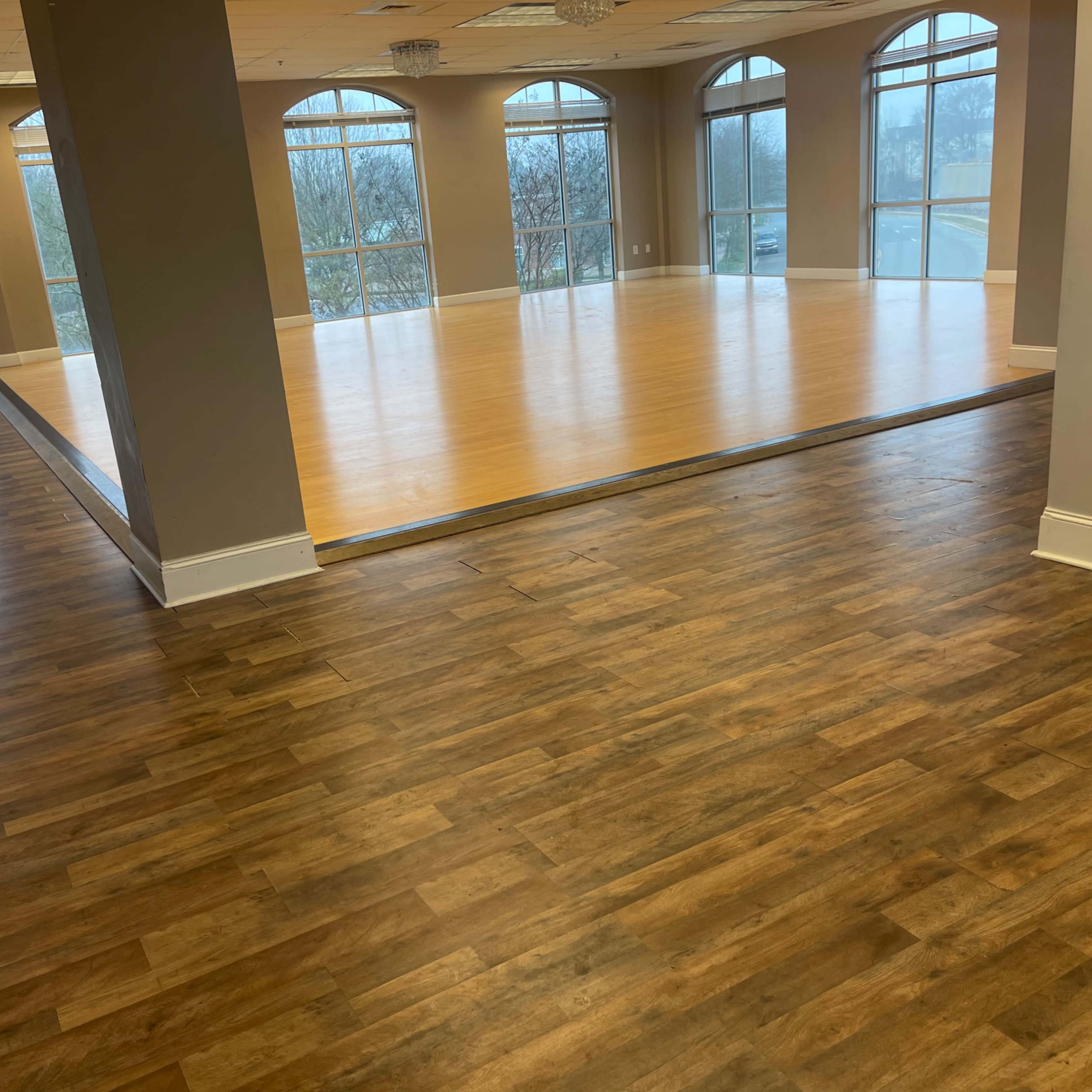 A spacious room with large windows, featuring a wooden floor and a smooth, light-colored dance area in the center.