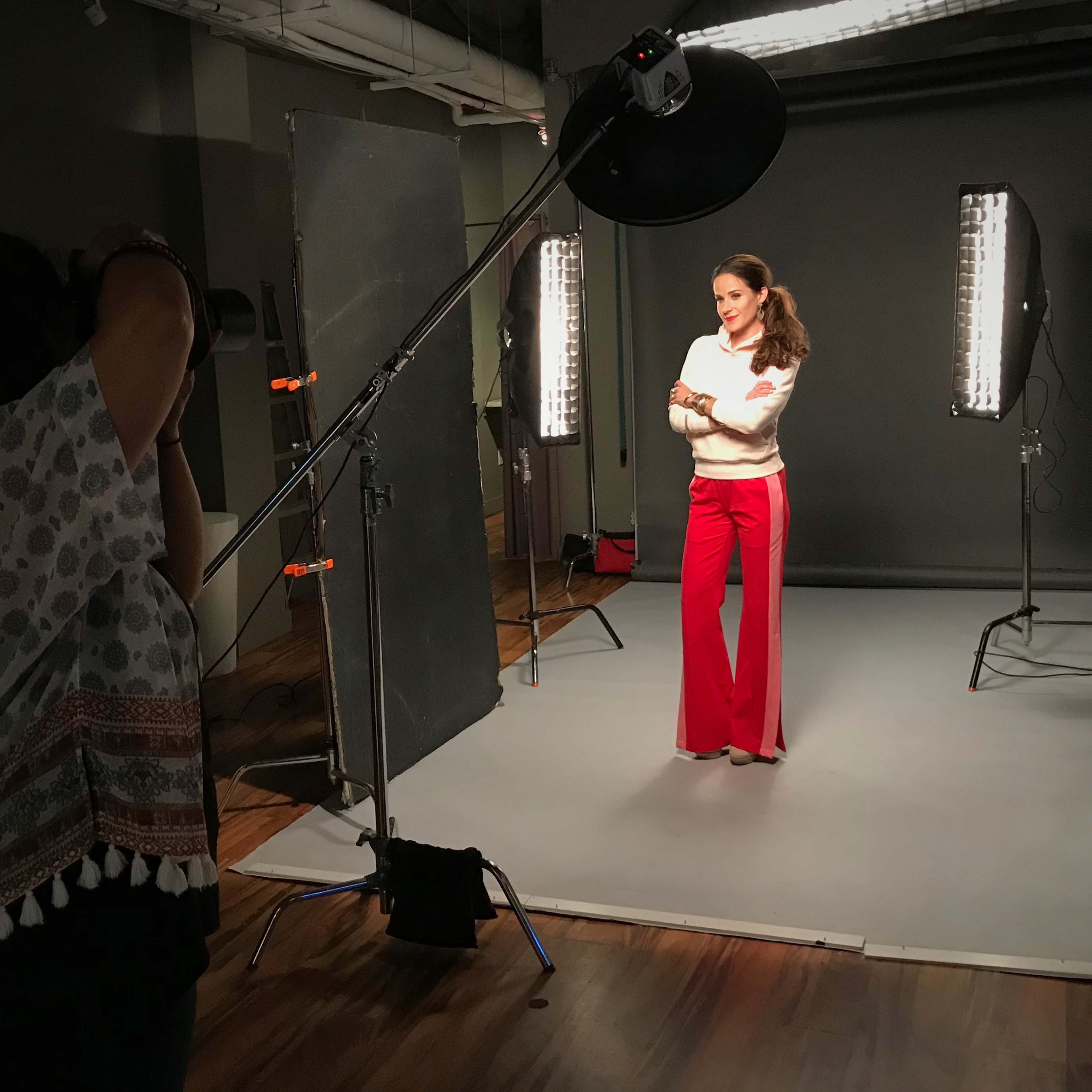 A photographer captures a subject posing in front of a gray backdrop with studio lighting equipment set up around them.