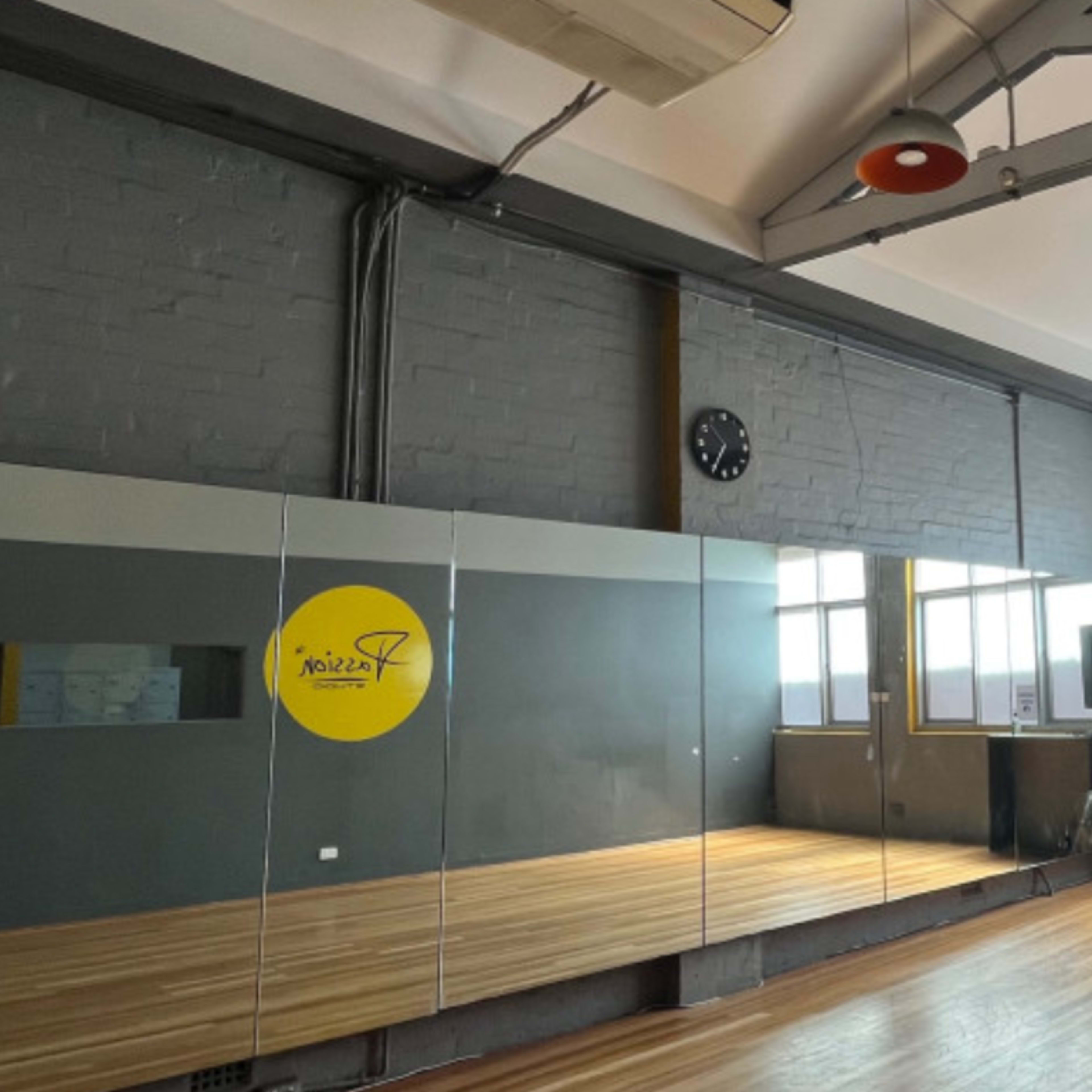 A spacious dance studio with wooden flooring, large windows, a wall mirror, and a wall clock.