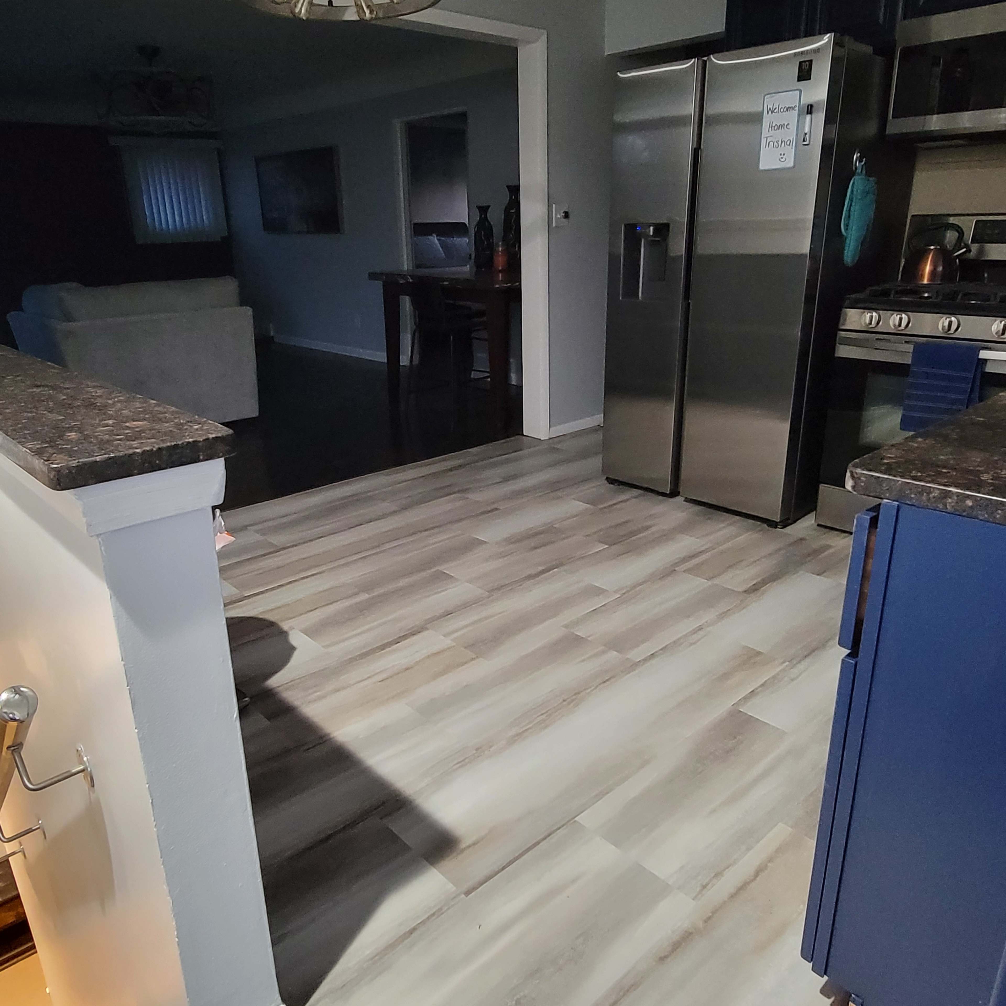 A kitchen area with stainless steel appliances, light-colored wooden flooring, and an adjoining living space visible in the background.