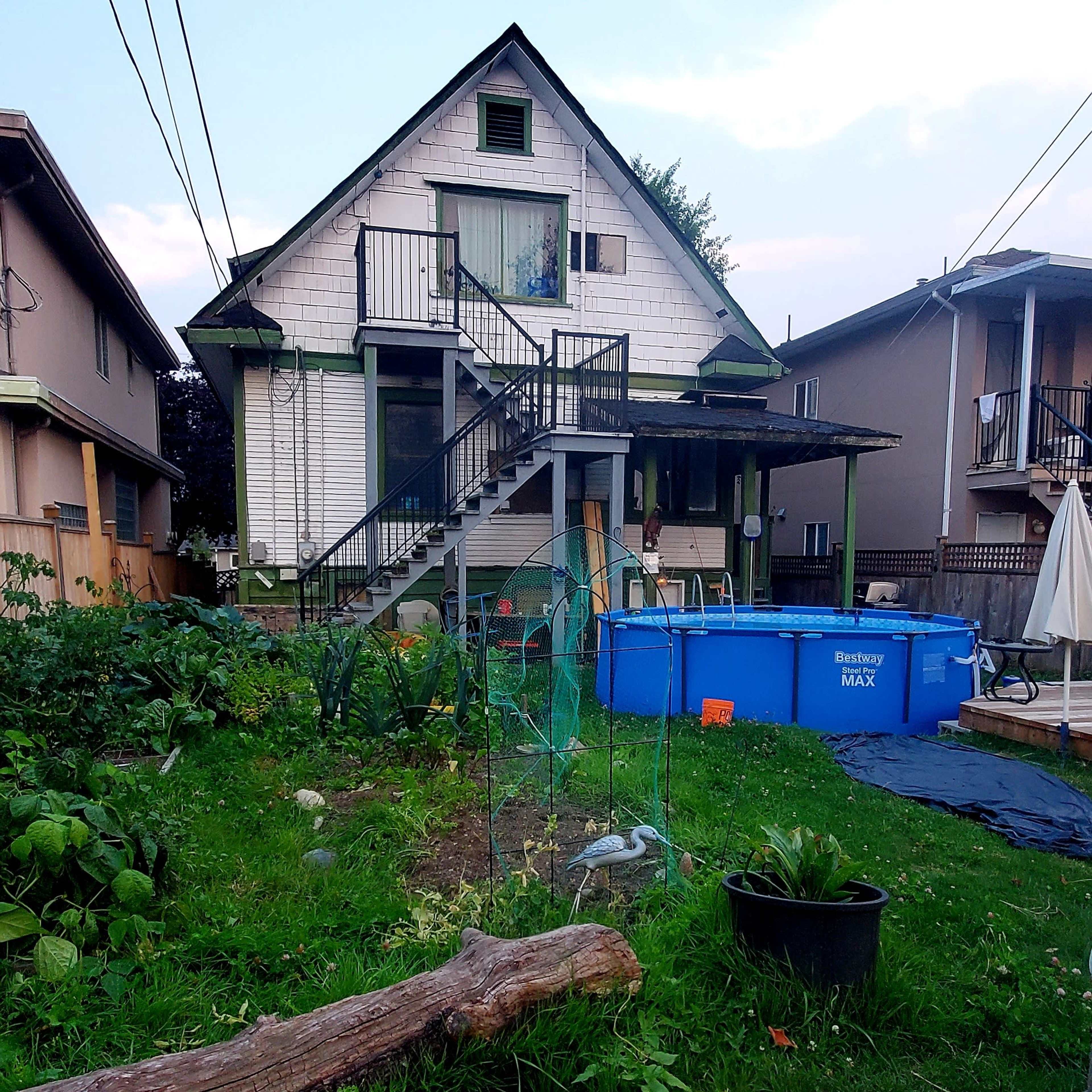 A two-story house with a sloped roof, stairs leading to a deck, and a pool in the backyard is surrounded by greenery and utility wires.