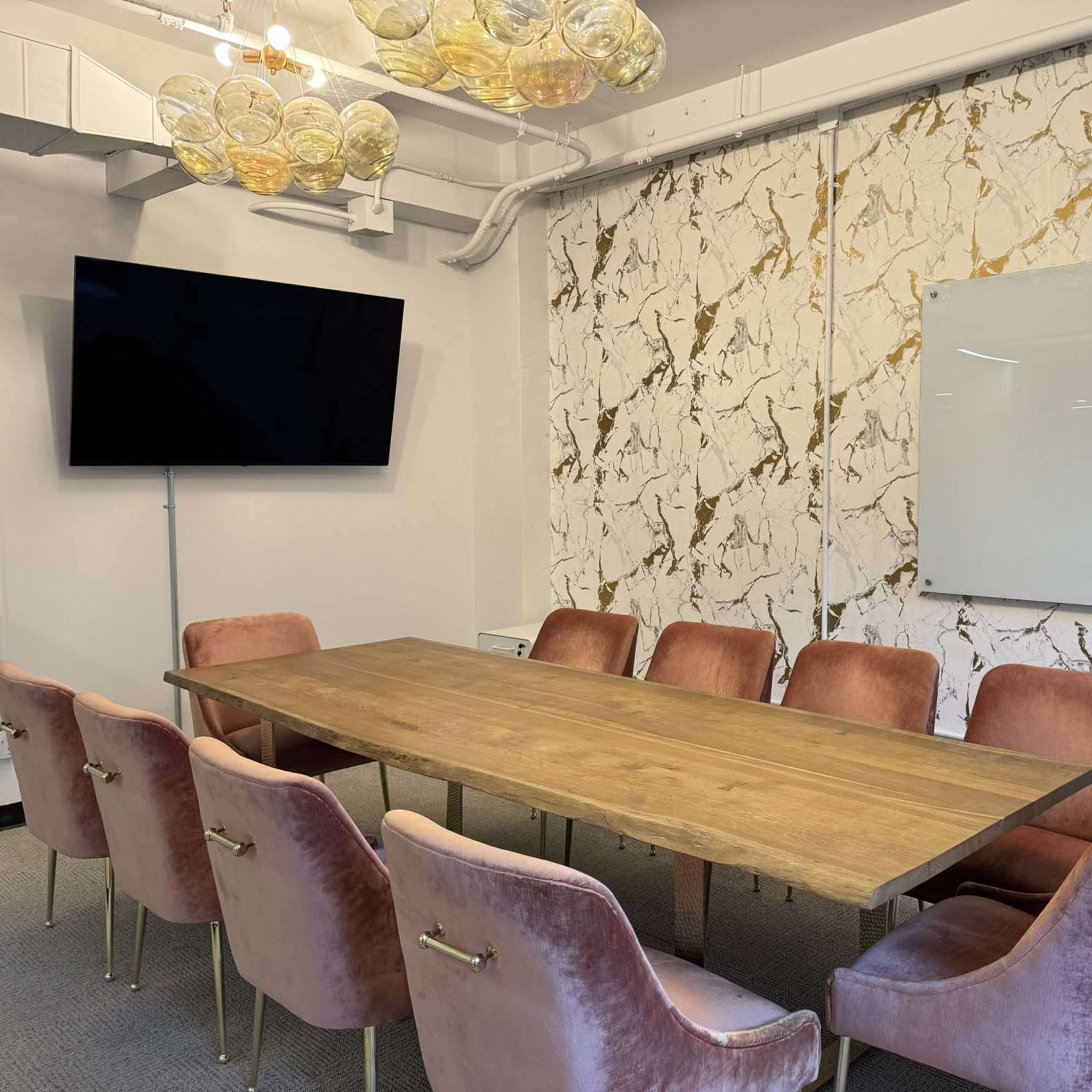A modern conference room featuring a long wooden table surrounded by pink upholstered chairs, a large television mounted on the wall, and a whiteboard beside a decorative marble-patterned wall.
