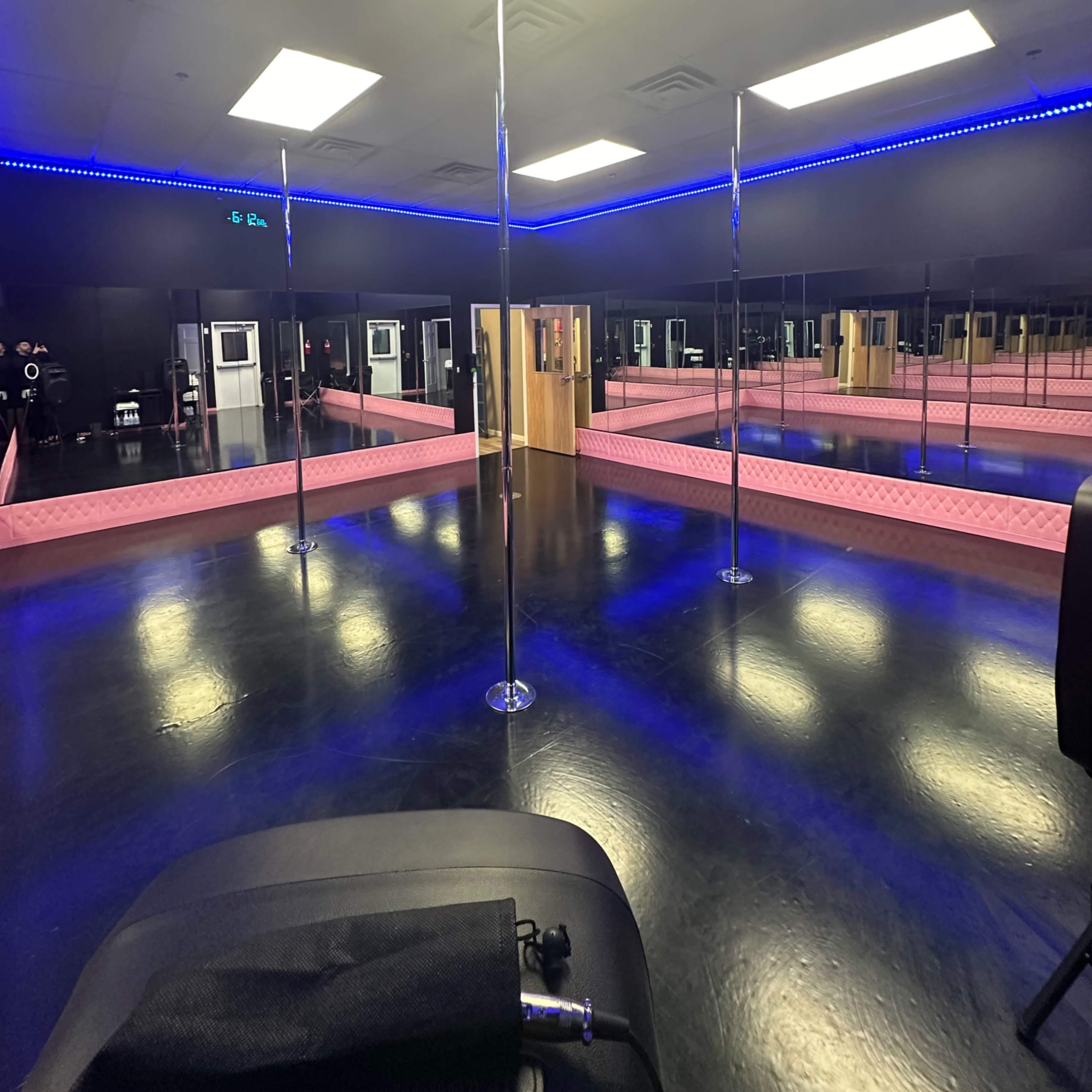 An empty dance studio featuring two vertical poles, large mirrors lining the walls, and blue LED lighting along the ceiling.