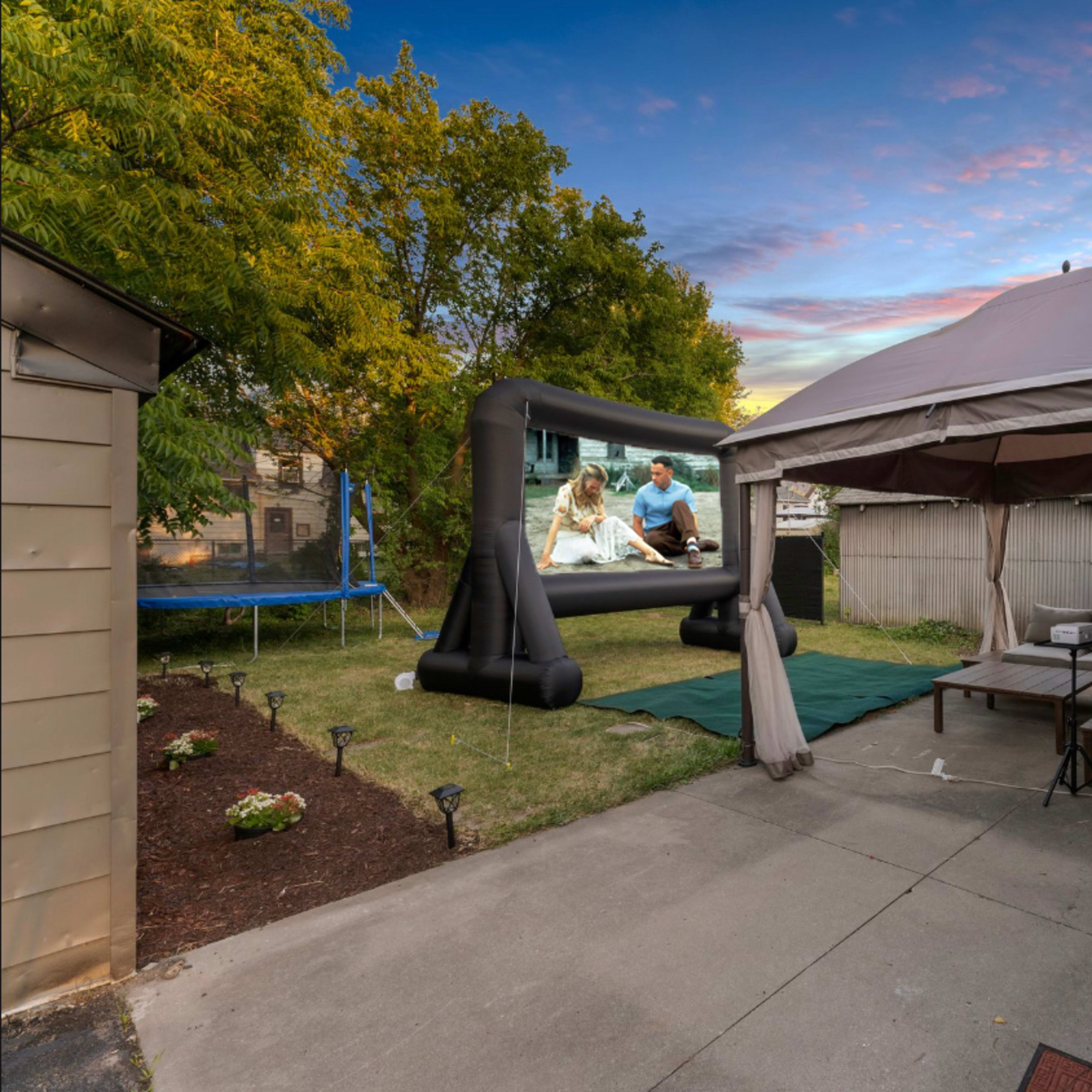 A backyard scene features a gazebo, outdoor seating, a grill, a trampoline, and a large inflatable screen displaying a video, set against a colorful sky.