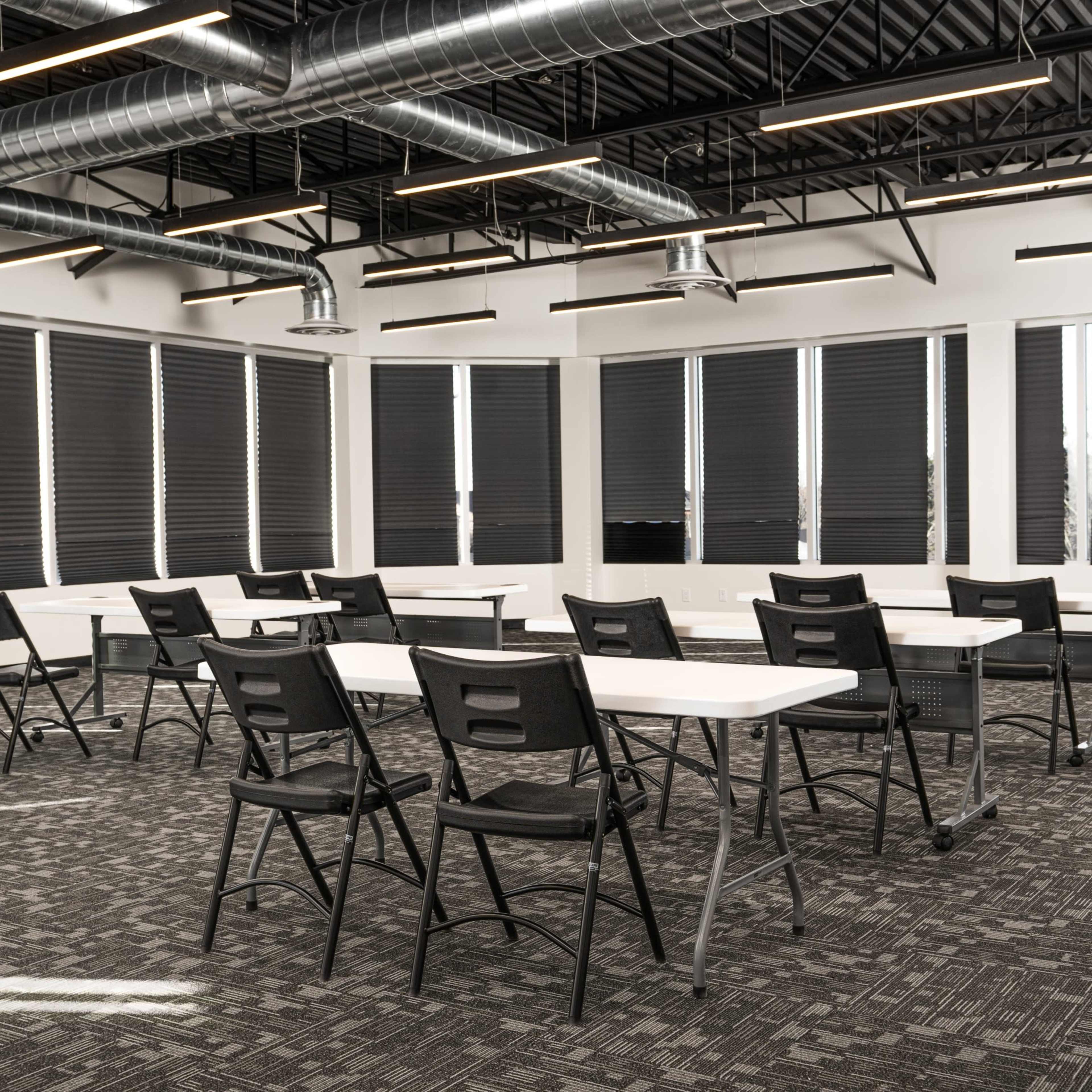A classroom features several rows of white tables and black chairs arranged on a carpeted floor, with large windows covered by retractable blinds.
