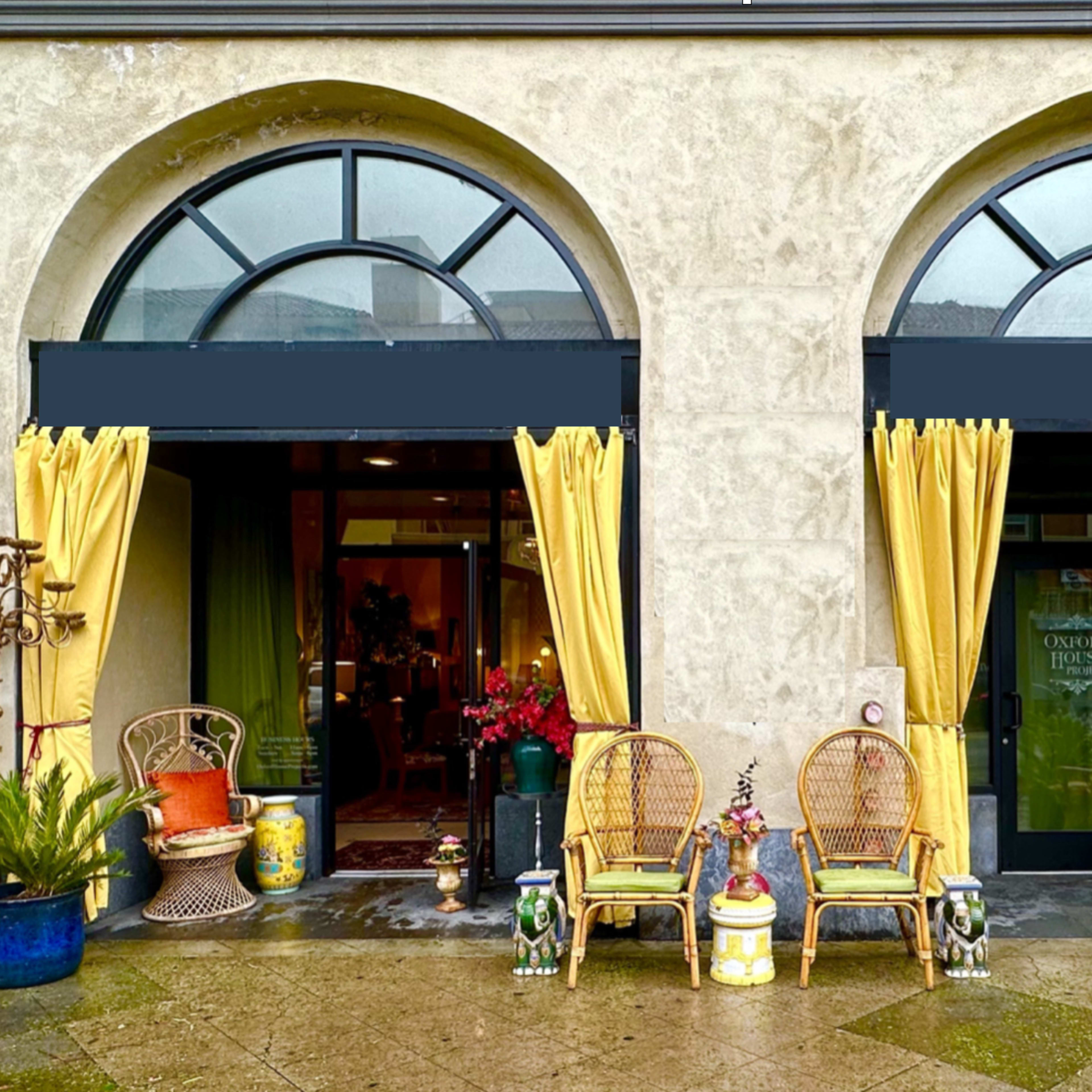 A building entrance with two arched doorways, each adorned with yellow curtains and flanked by potted plants and wicker chairs.