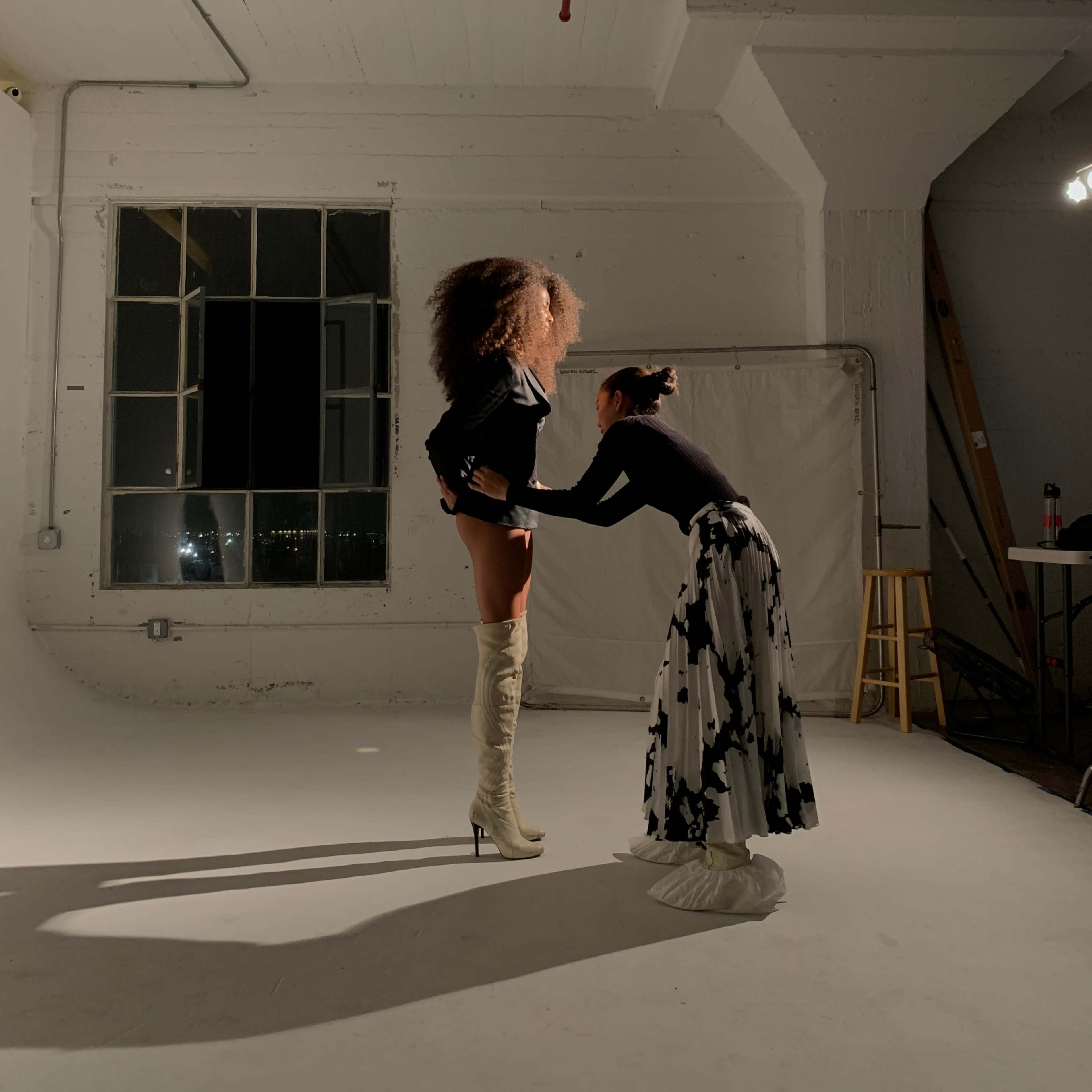 A model in knee-high boots poses as a stylist adjusts the outfit in a bright studio with large windows.