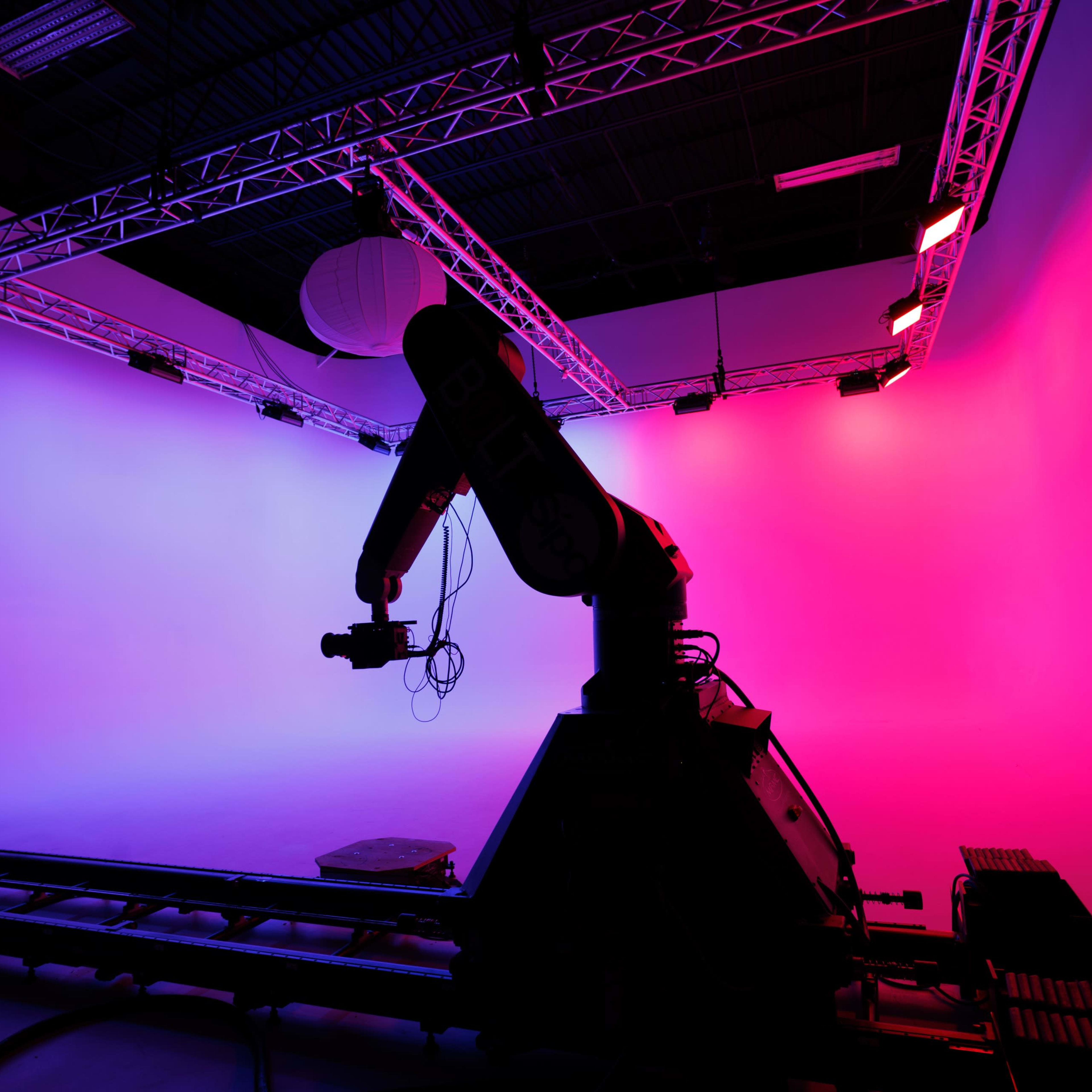 A robotic arm is positioned in a studio space with colorful lighting against a backdrop of gradient pink and blue.