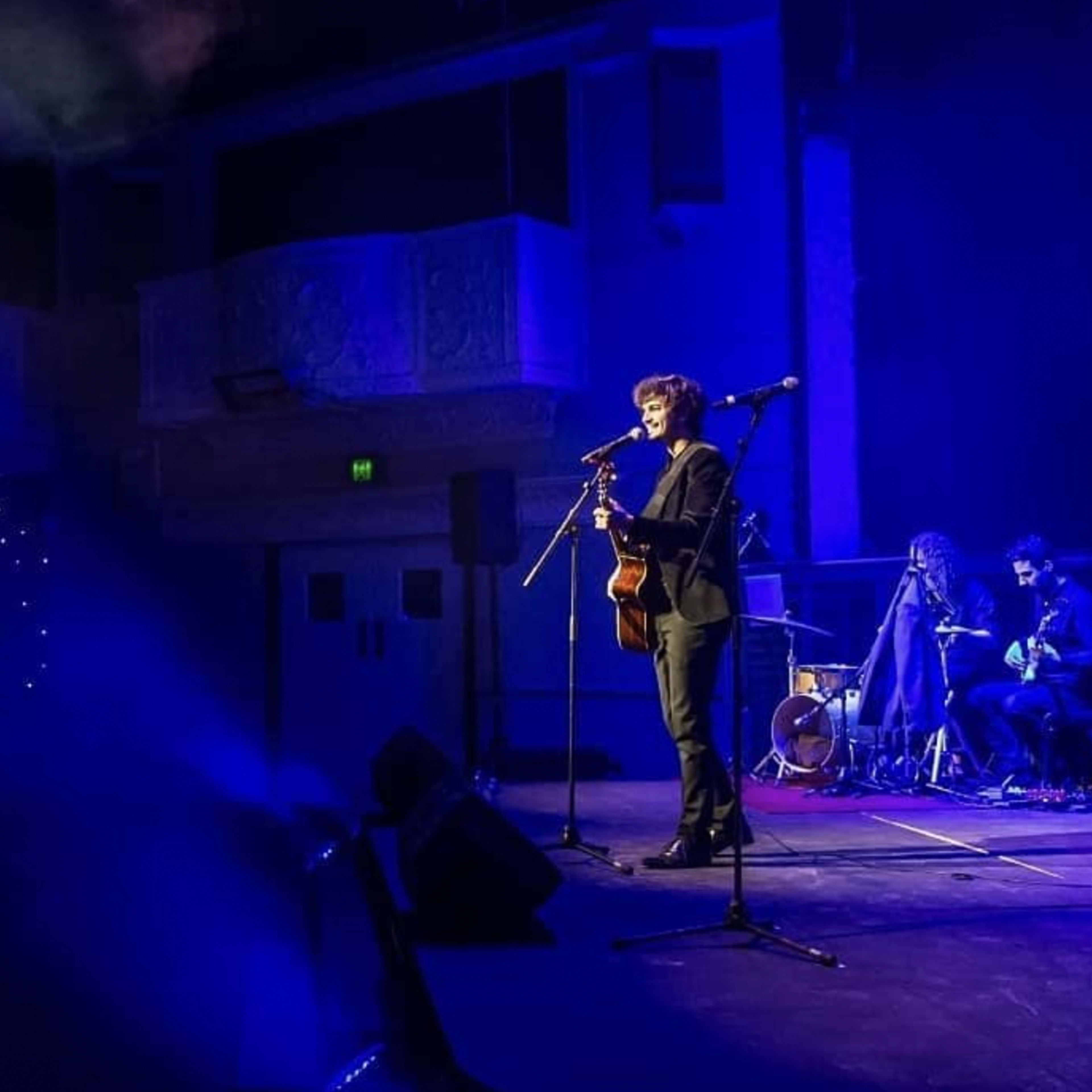 A musician performs on stage with a guitar while a band accompanies him in a dimly lit venue.