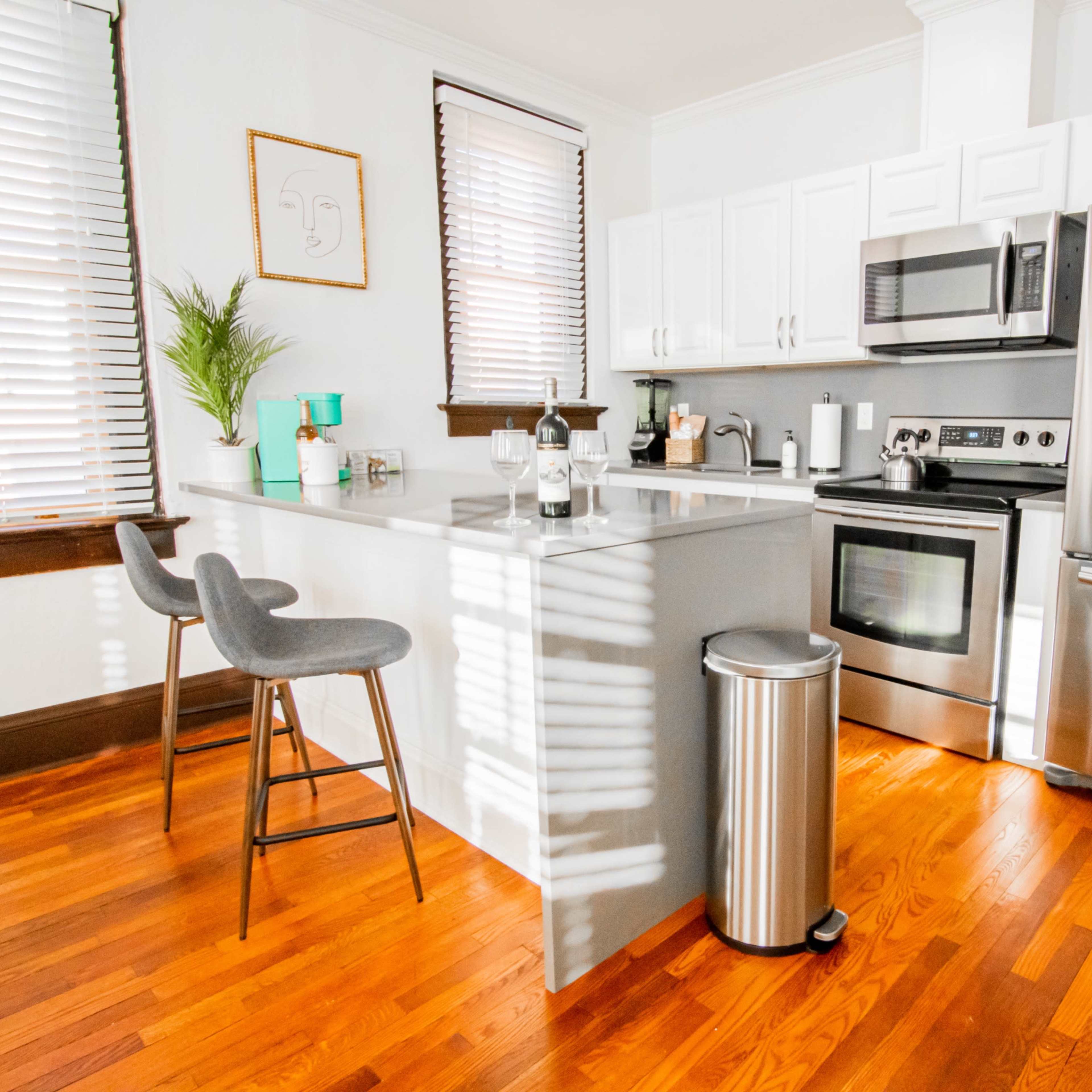 A modern kitchen with stainless steel appliances, a gray countertop island, and wooden flooring, featuring two bar stools and a window with blinds.
