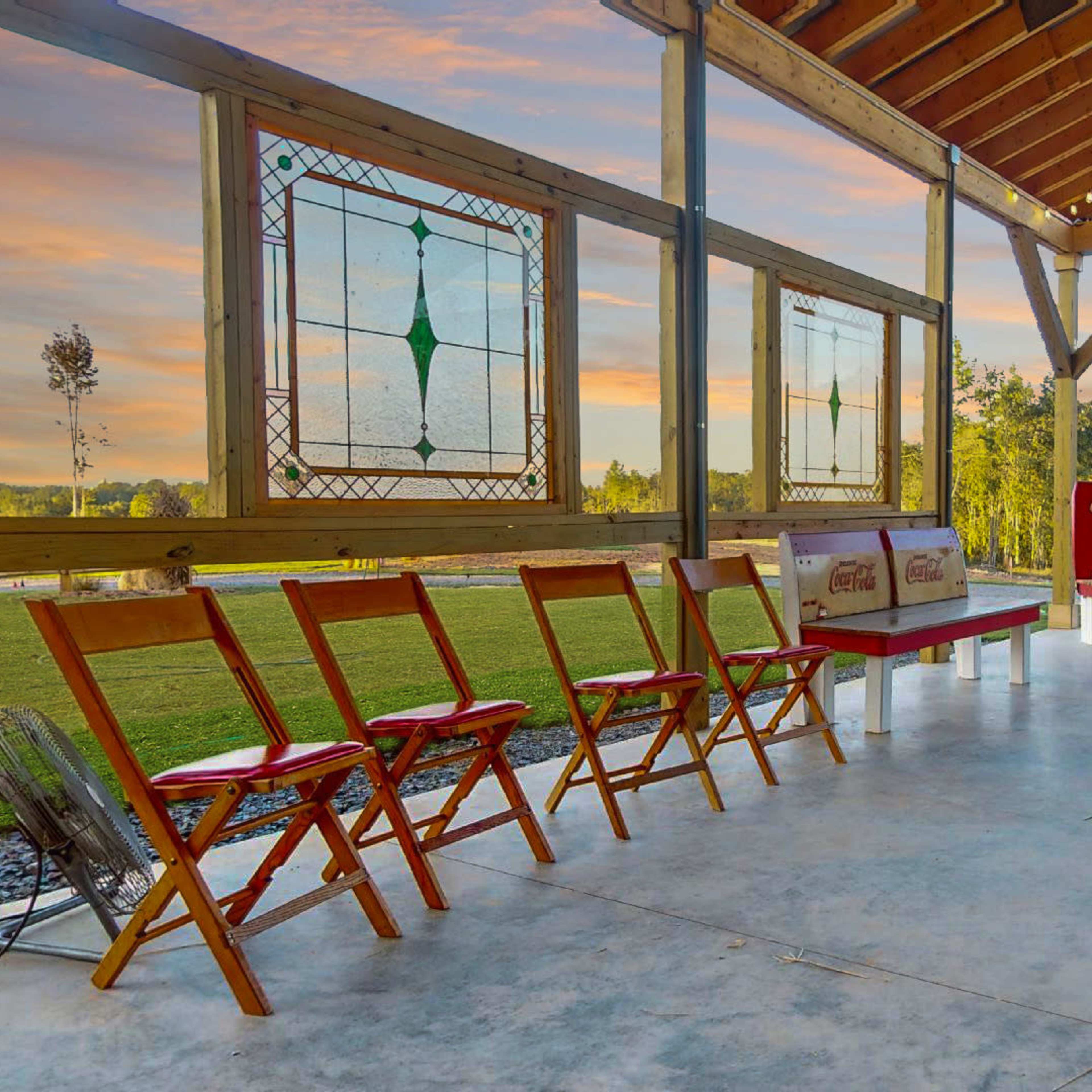 A spacious outdoor pavilion with wooden beams, colorful stained glass windows, and several folding chairs arranged around a circular area near a grassy field.