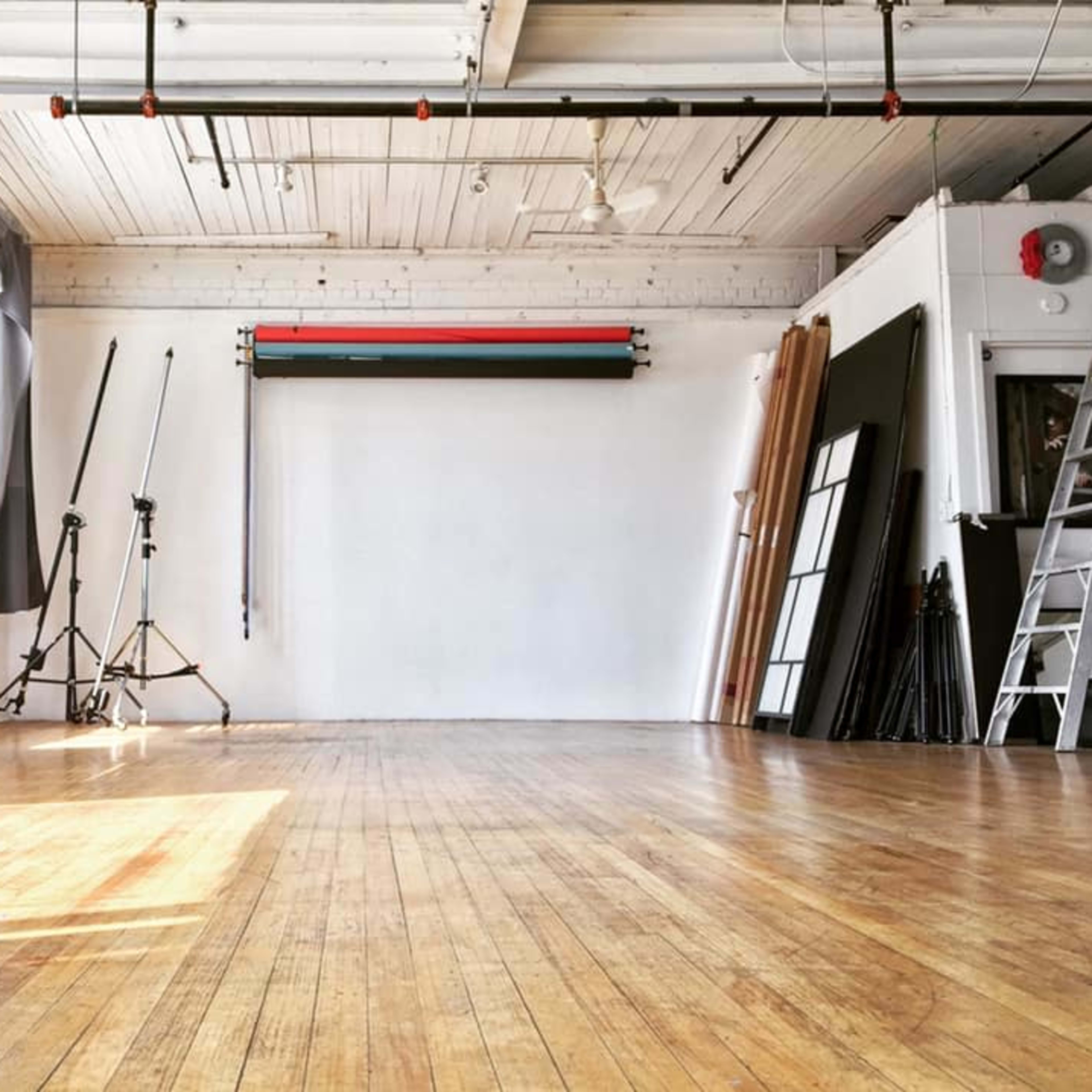 A spacious, empty photography studio with wooden floors, tall windows, and various equipment along the walls.