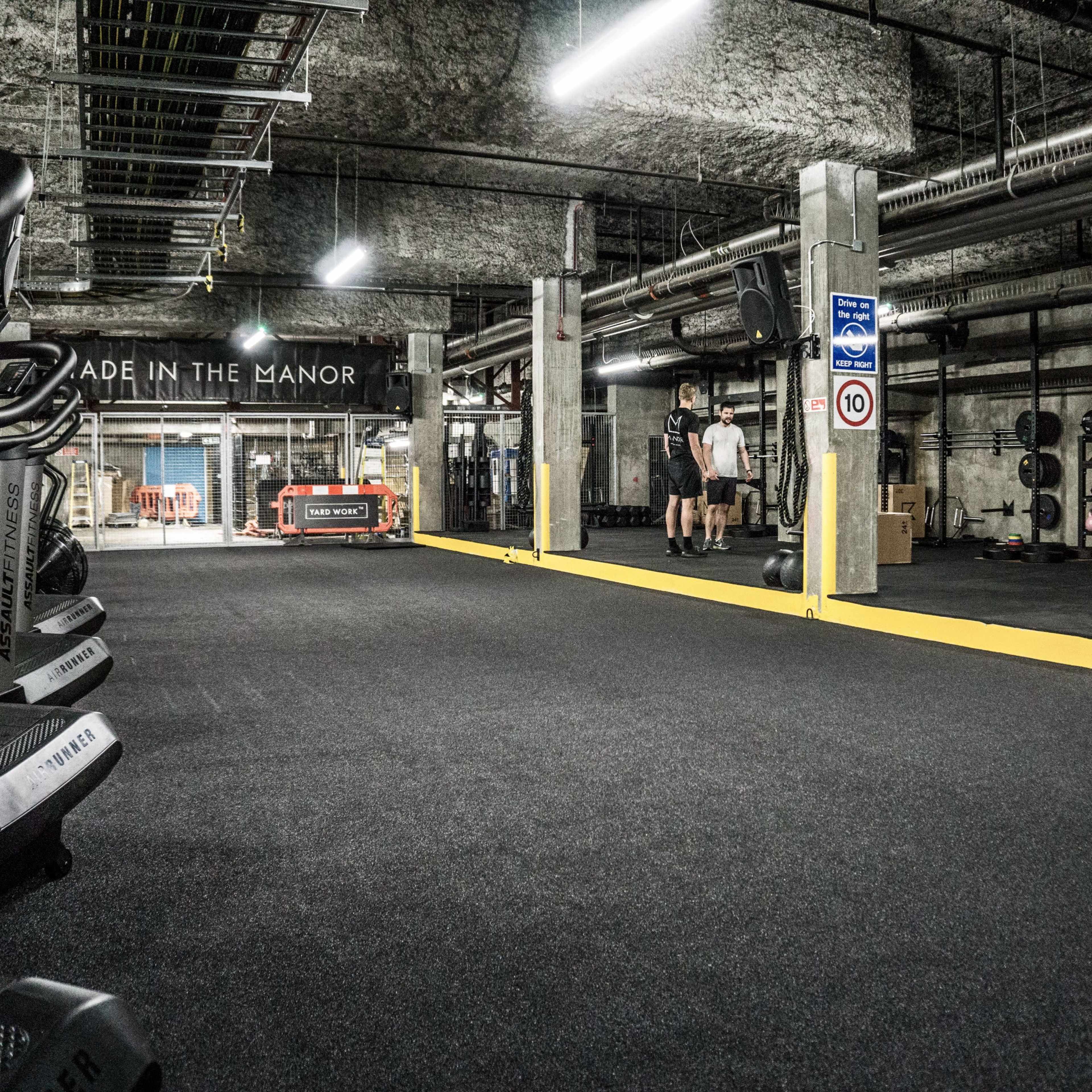 A spacious underground gym features various exercise equipment, including treadmills and weights, with two individuals engaging in a workout.