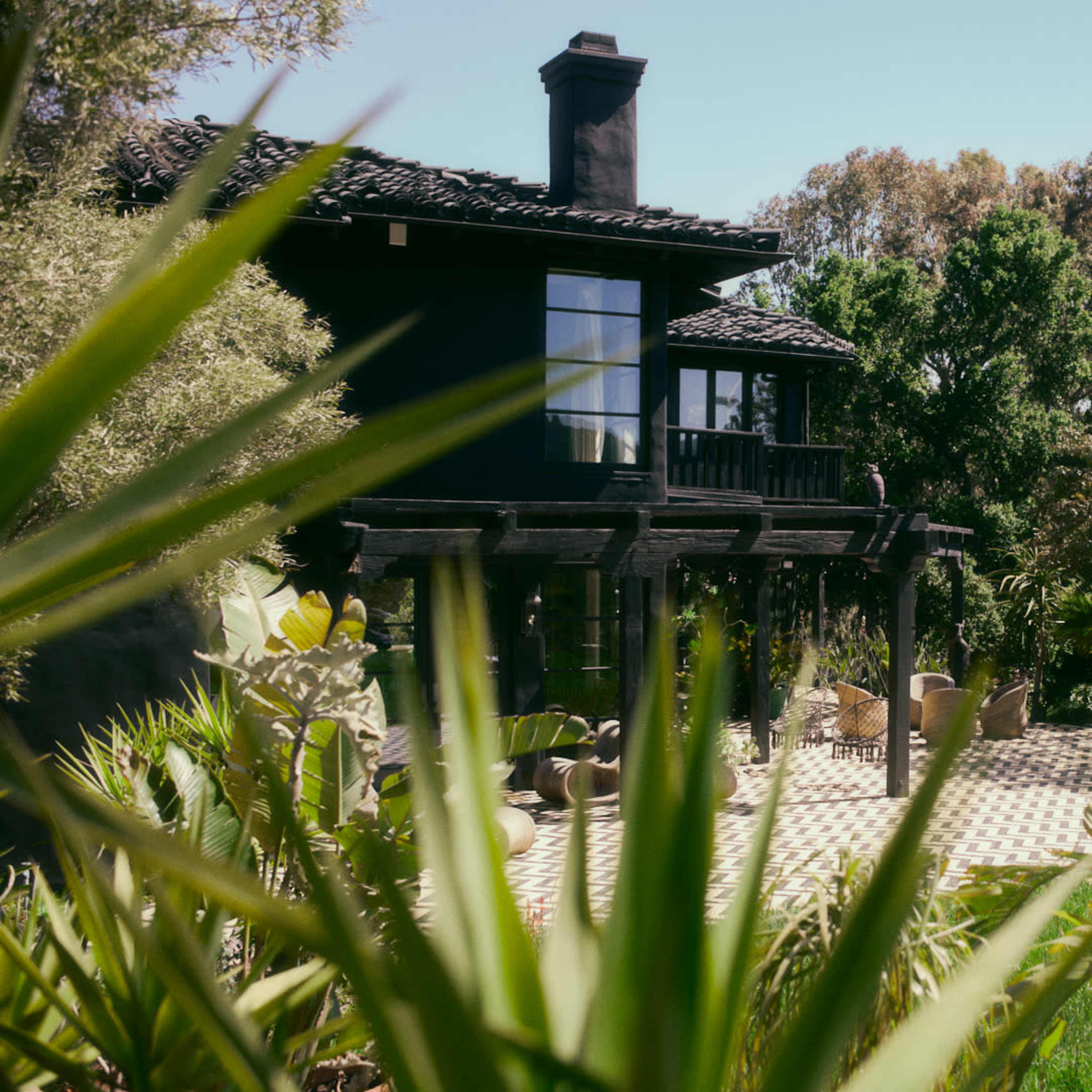 A black house with a tiled patio is surrounded by lush greenery and seated areas.