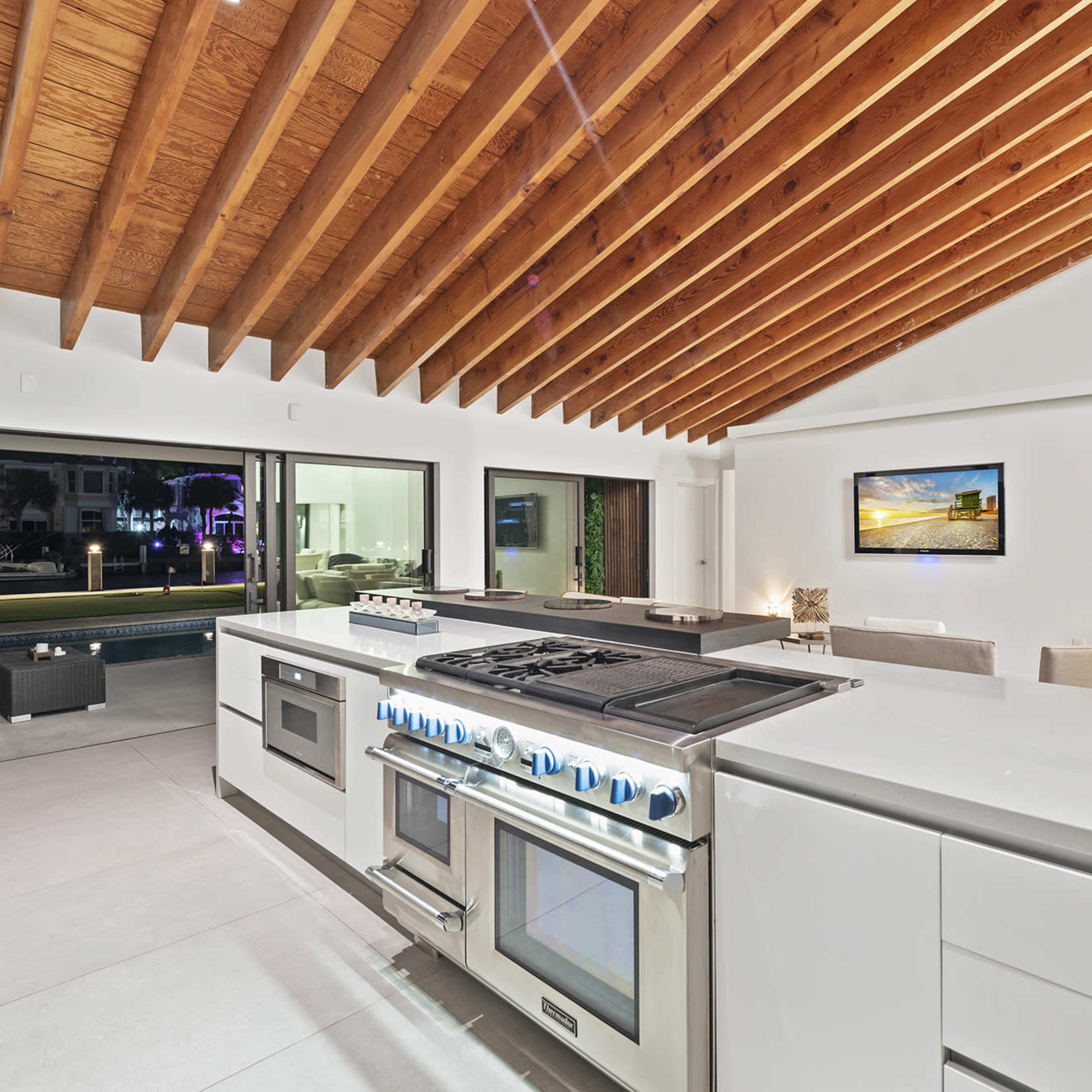 A modern kitchen features a large central island with a professional-grade stove and expansive windows overlooking a patio and pool area.