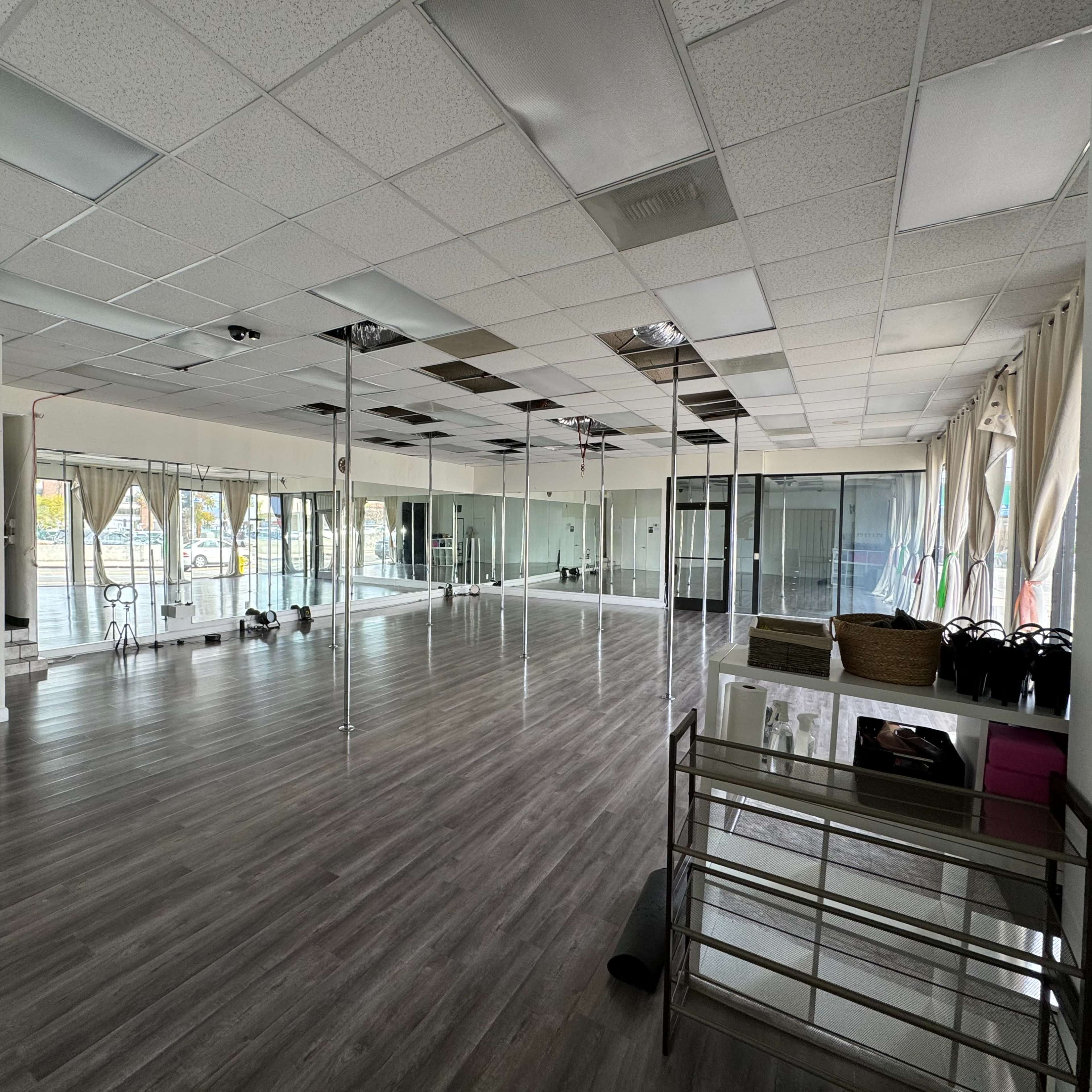 An empty dance studio with mirrors lining one wall, wooden flooring, and various exercise equipment in the corner.