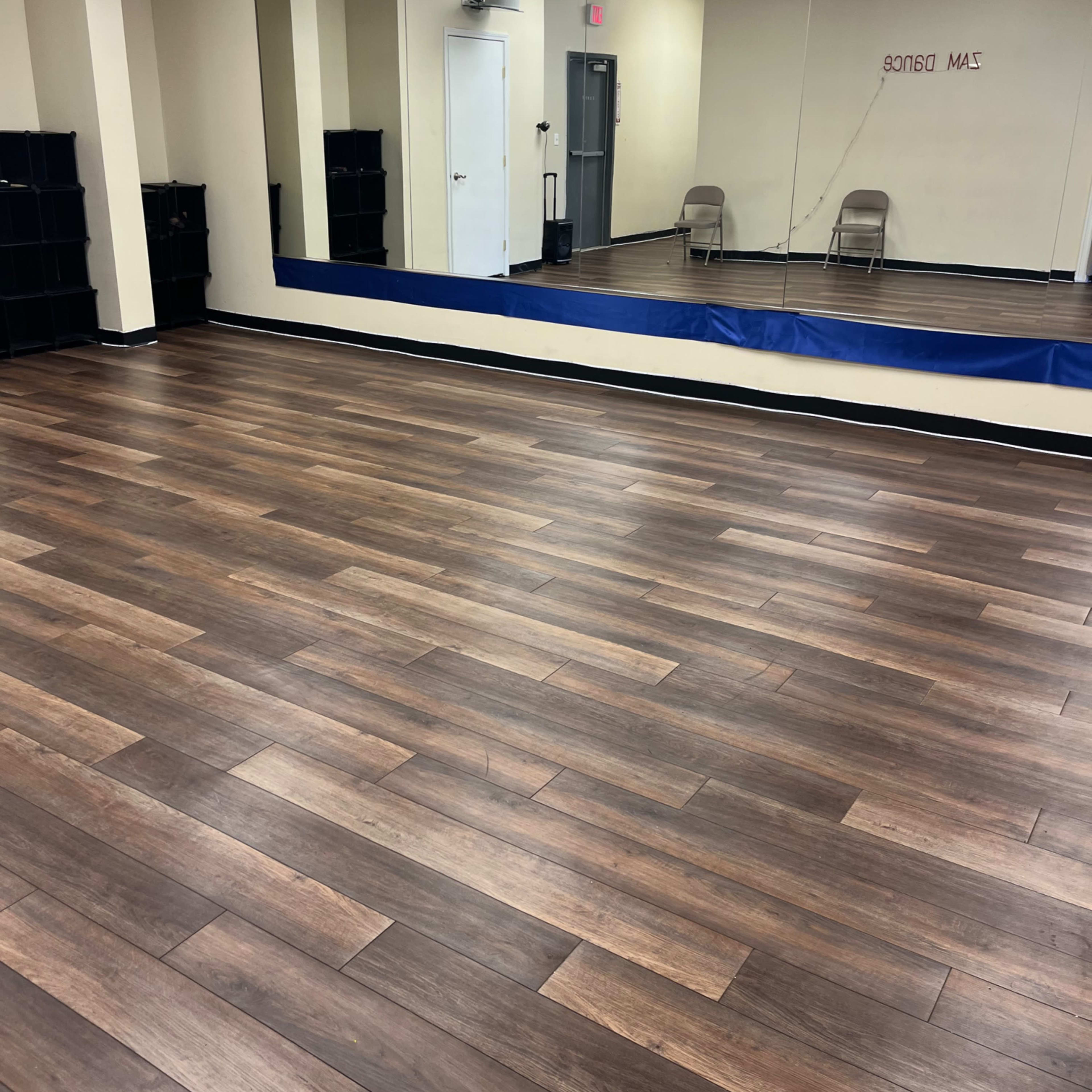 An empty dance studio with wooden floors, a large mirror on one wall, and two folding chairs in the background.