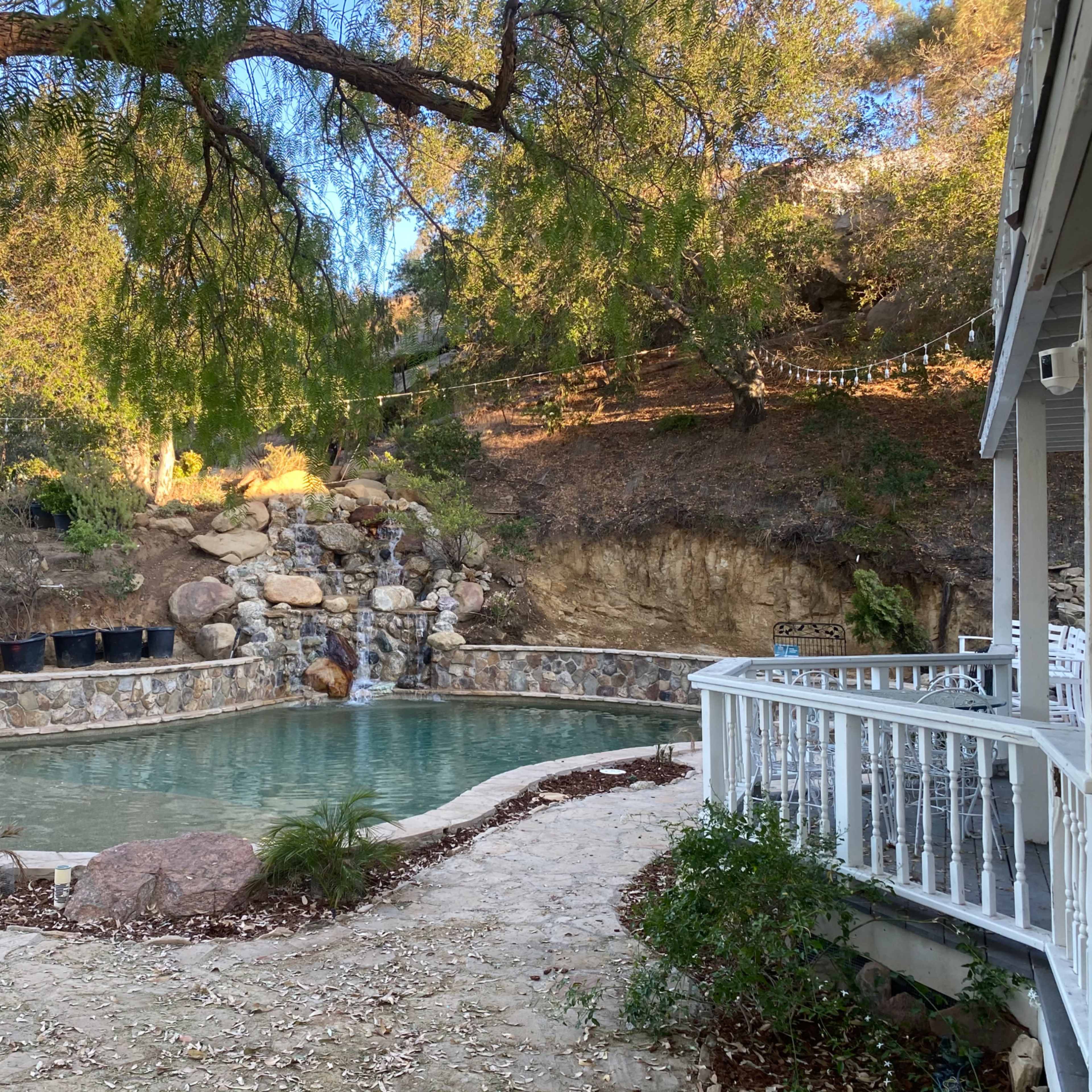 A stone-bordered swimming pool is situated in a rocky landscape with trees and a small waterfall in the background.