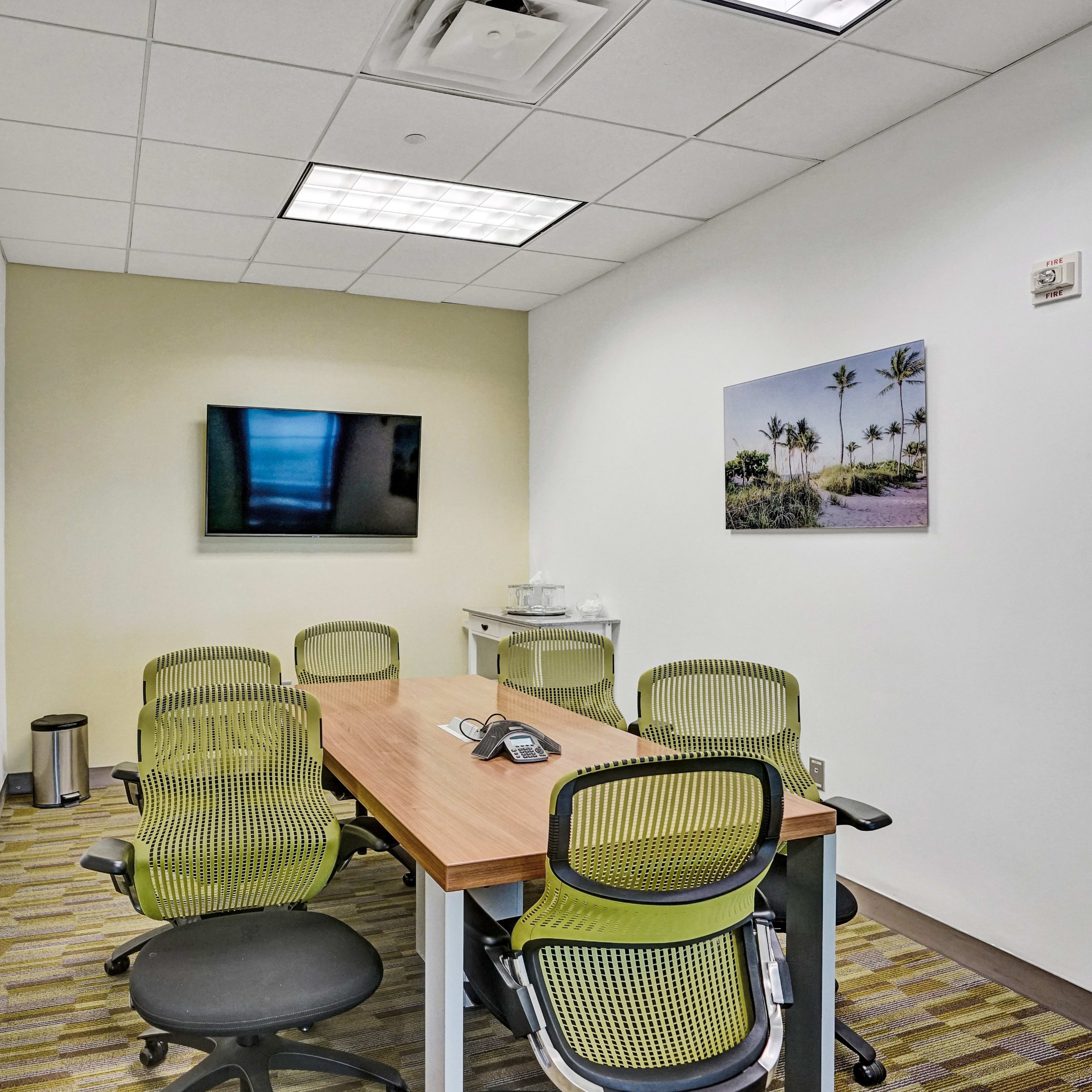 A meeting room with a rectangular table surrounded by green office chairs, a television mounted on the wall, and a palm tree photograph framed on another wall.
