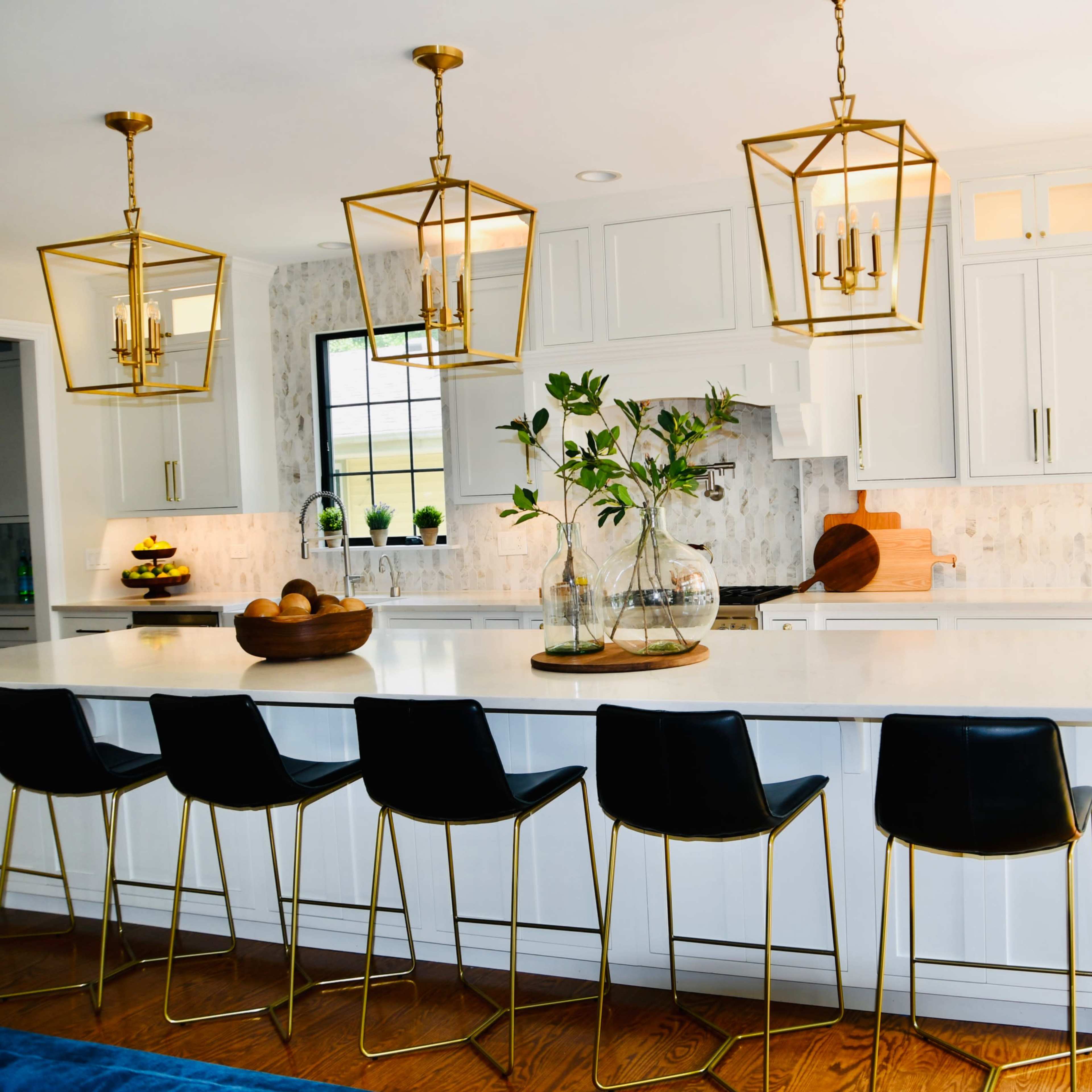 A modern kitchen features a large island with black bar stools, gold pendant lights, and white cabinets, complemented by a light-toned backsplash and stainless steel appliances.