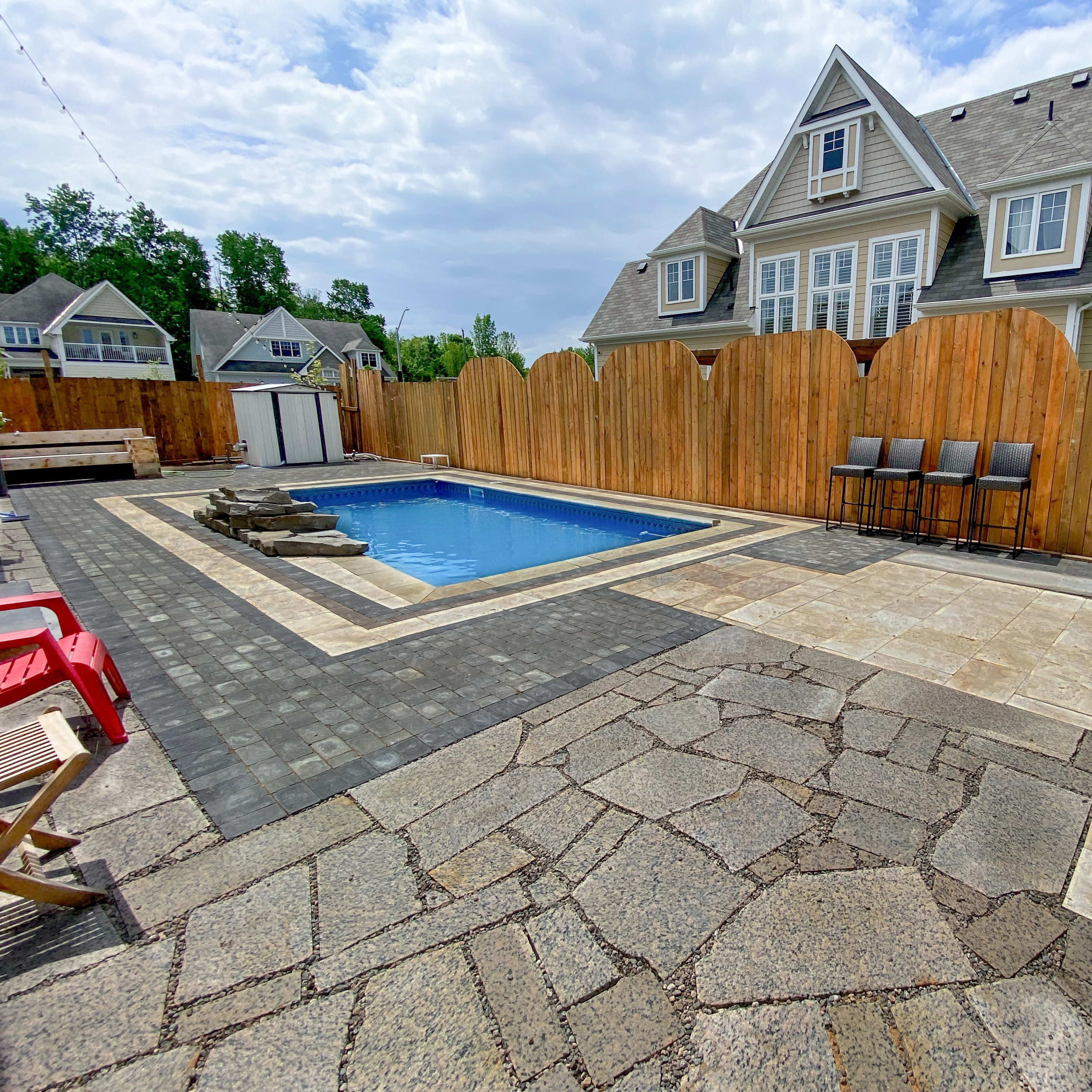 A rectangular swimming pool surrounded by a stone patio, with a wooden fence and a house in the background.