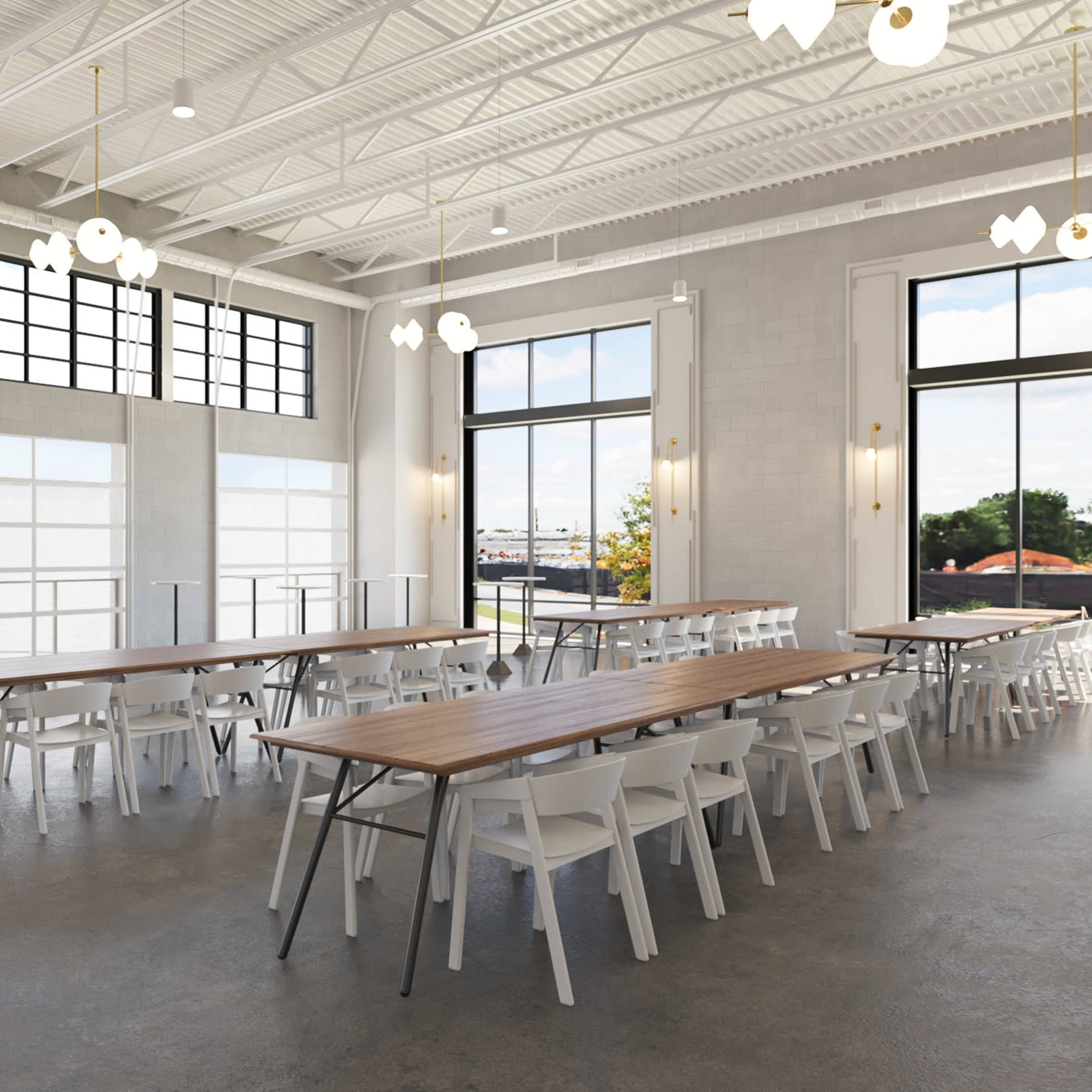 A spacious event hall with long wooden tables and white chairs, large windows allowing natural light, and a setup area with a decorative wall.