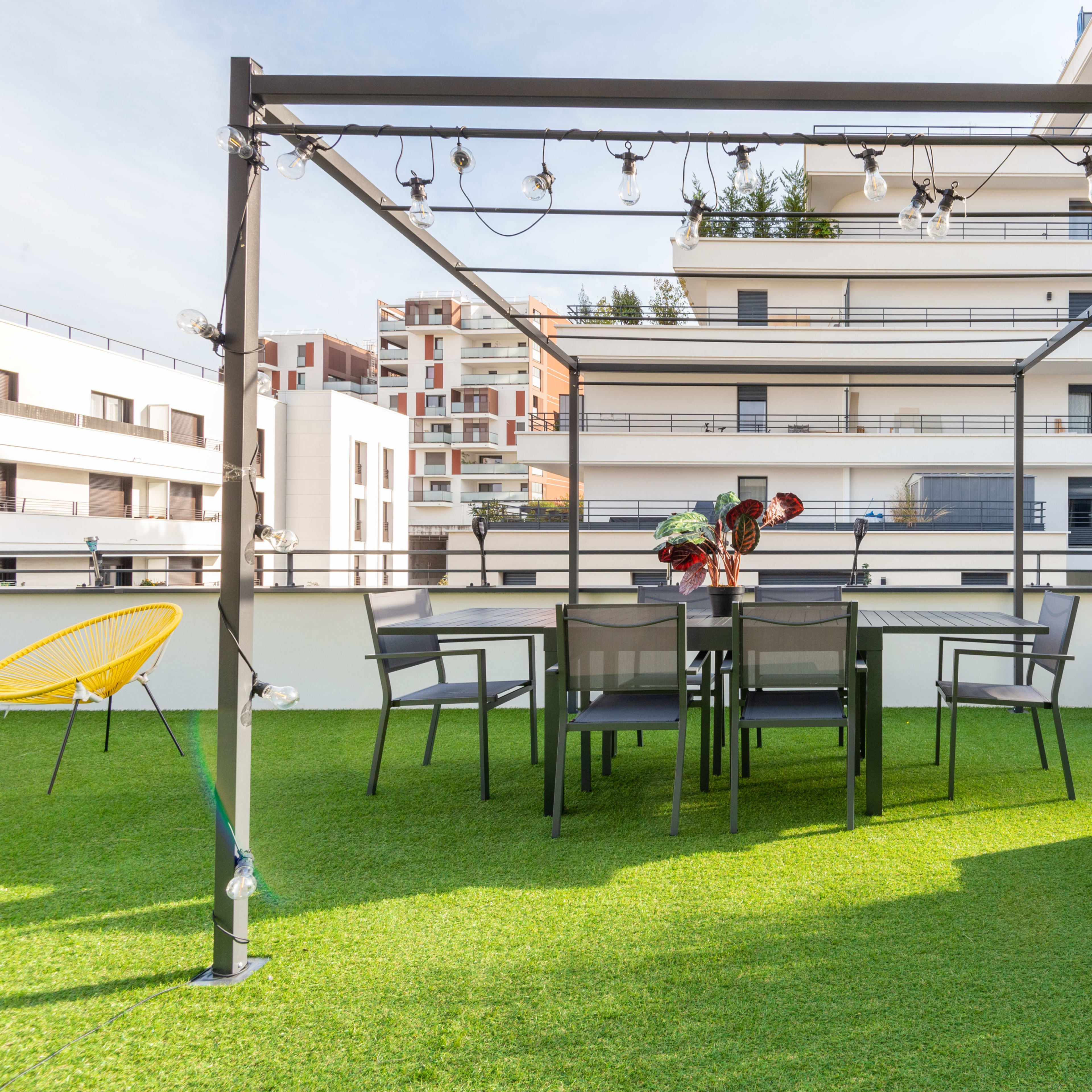 A patio area featuring a dining table and chairs under a metal pergola with string lights, surrounded by green artificial grass.