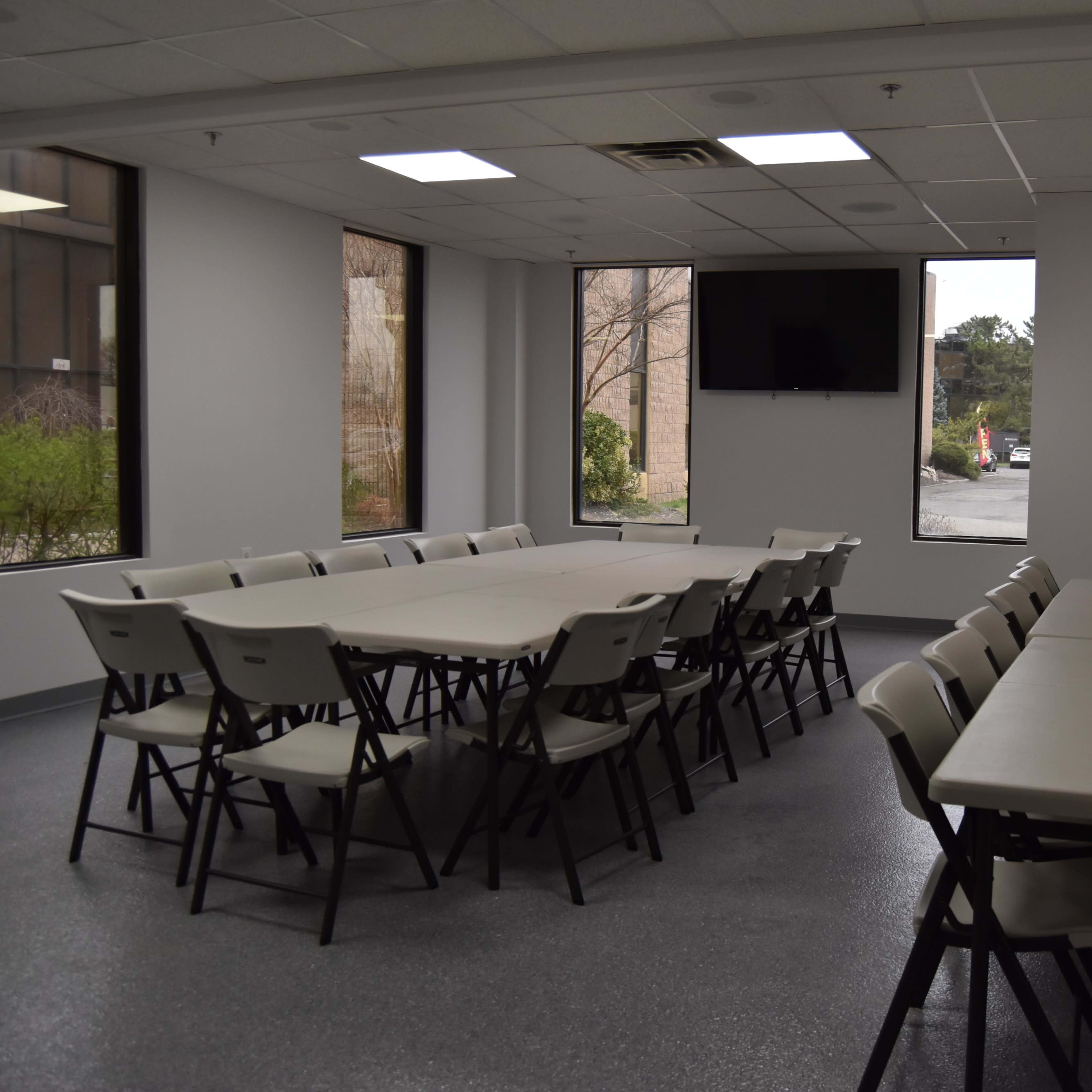 A meeting room features a large rectangular table surrounded by chairs, with large windows on two sides and a monitor mounted on the wall.