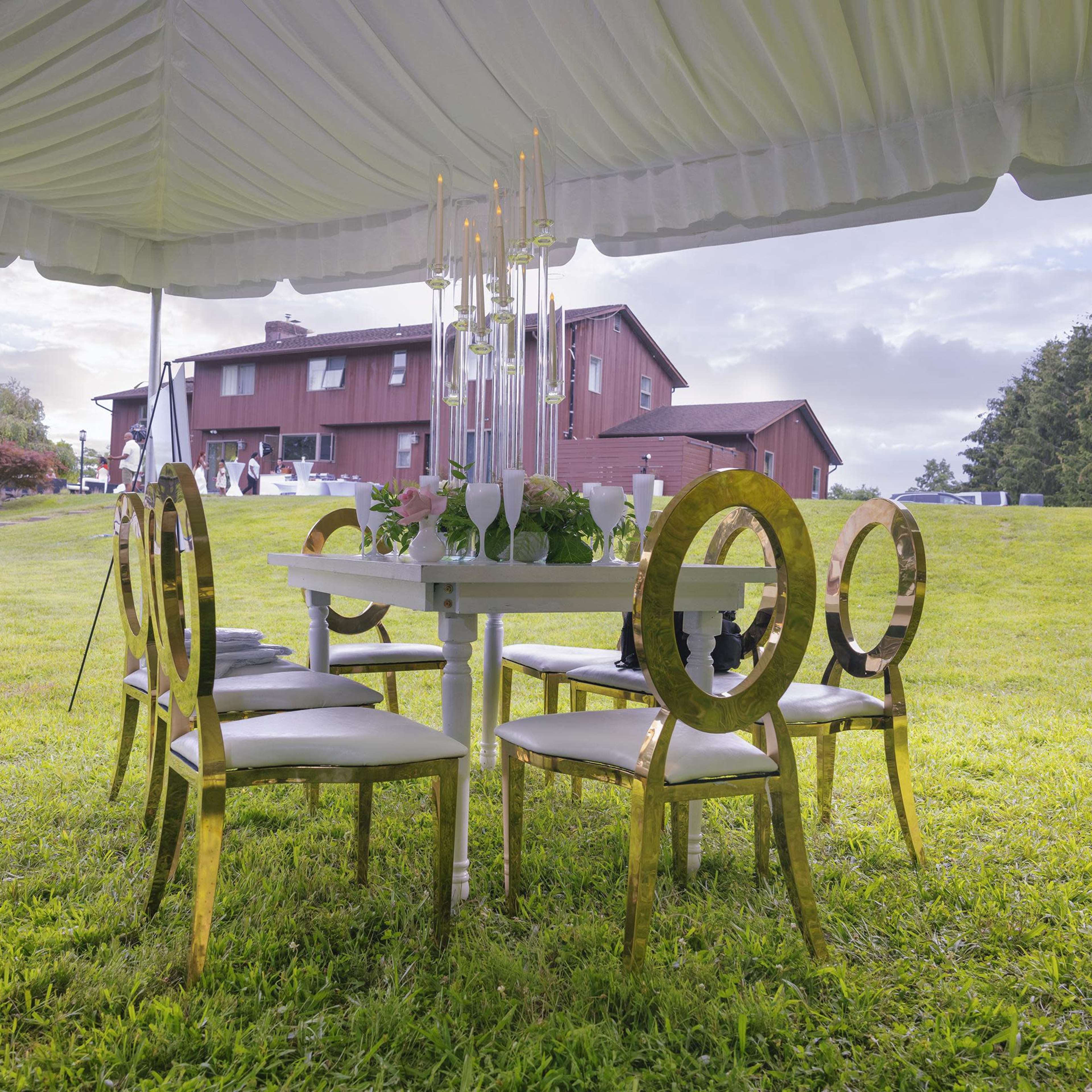 A round table with gold-accented chairs is set up under a tent in a grassy area, with a house visible in the background.
