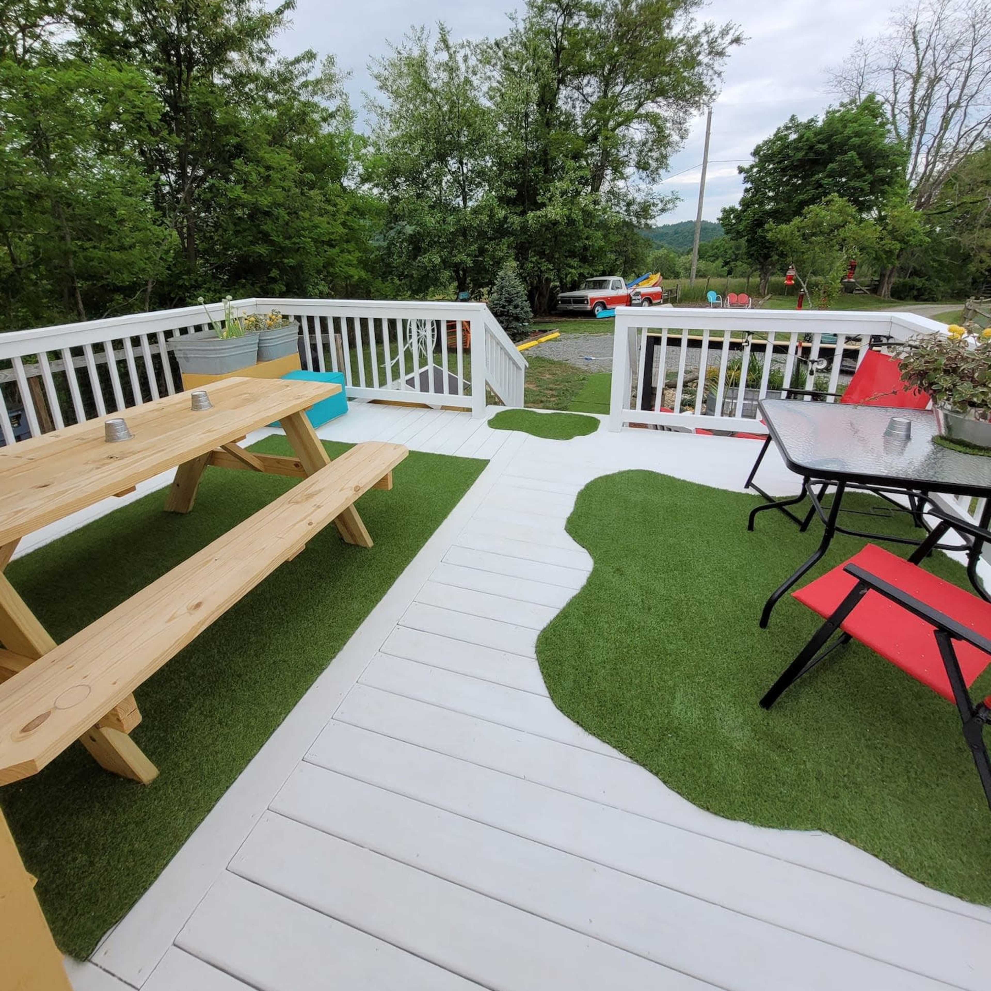 A deck with wooden picnic tables and black and red outdoor chairs, surrounded by green grass and trees.