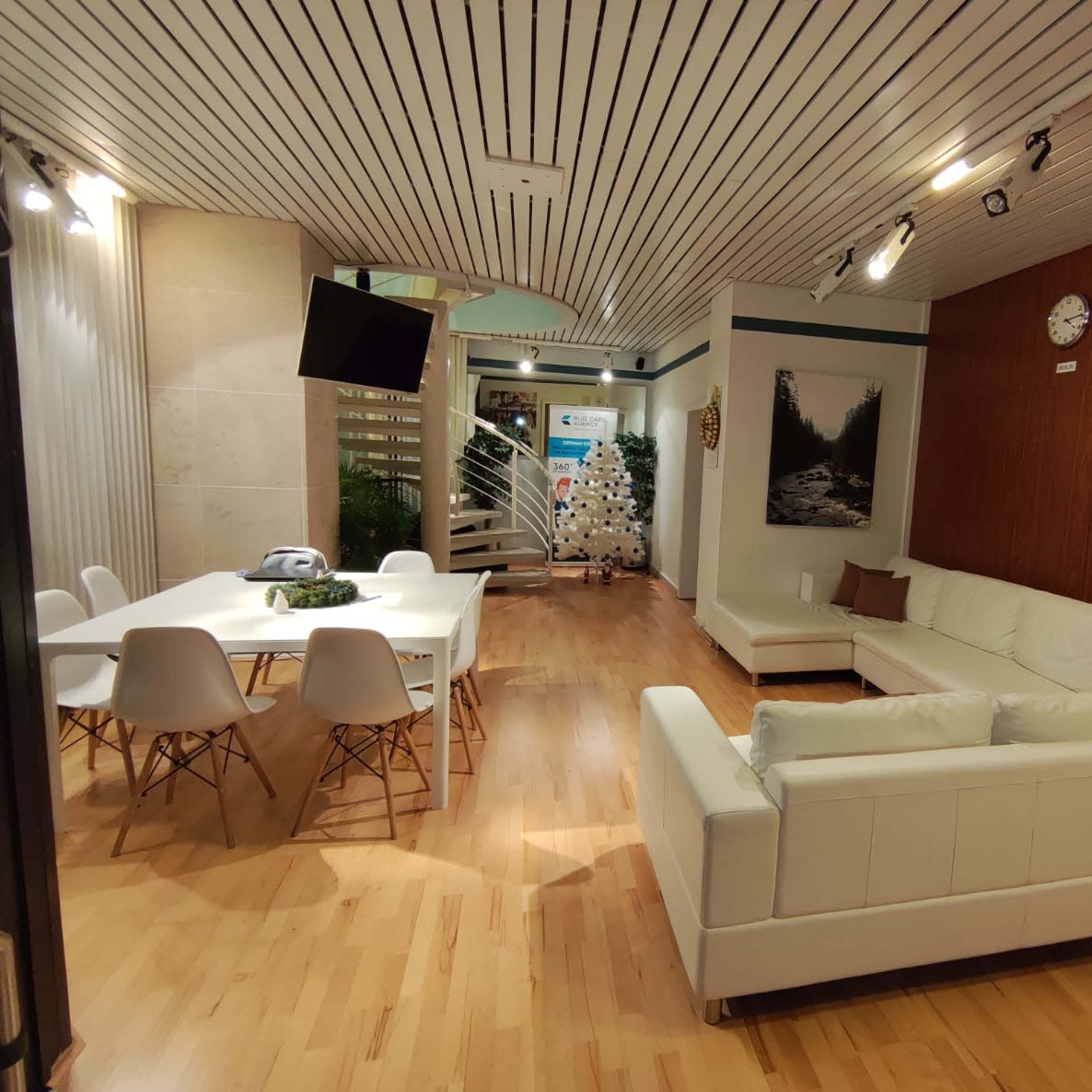 A modern office interior with a white sofa, a dining table, and a TV on the wall, featuring wooden and light-colored floors.