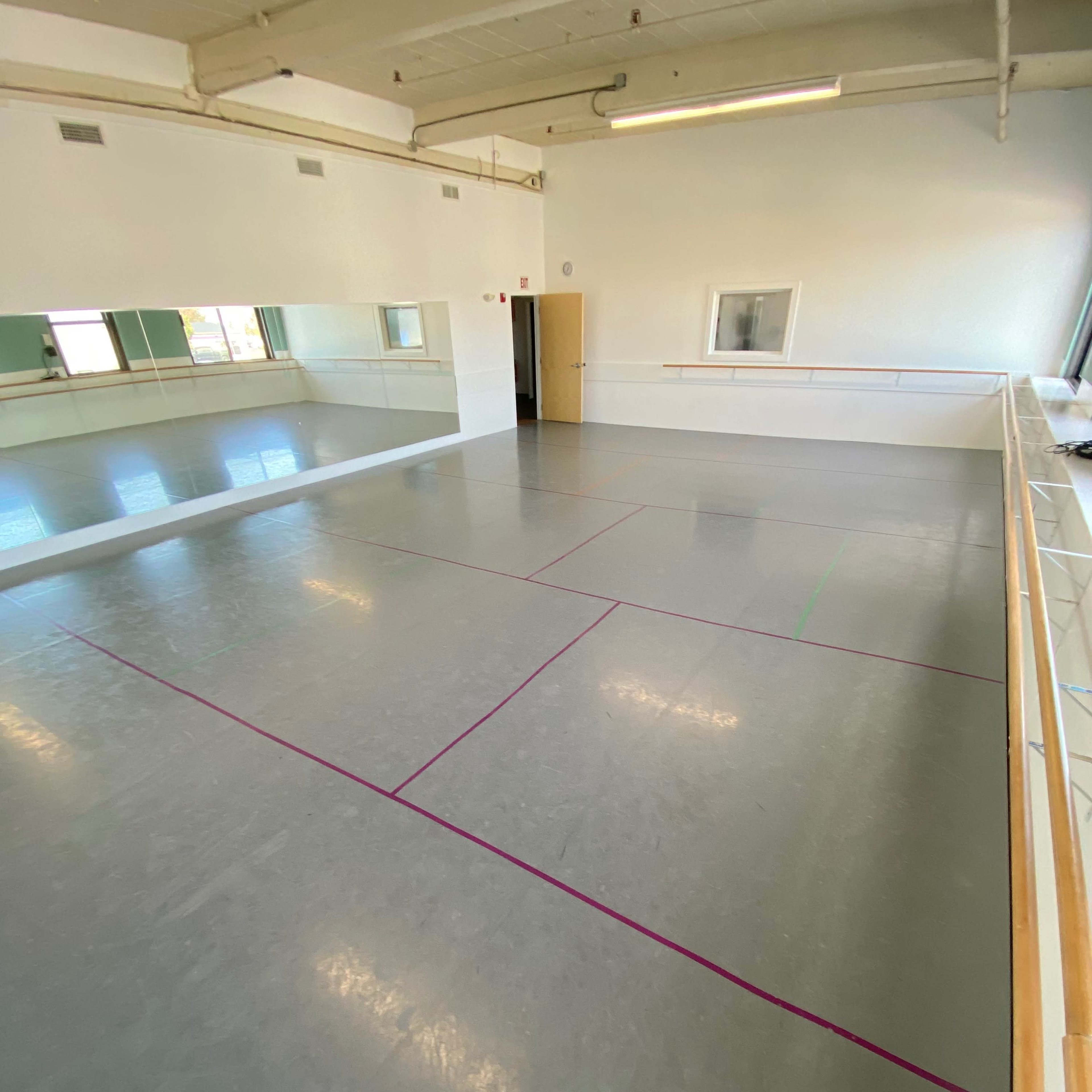 A spacious dance studio with a gray floor marked by colored lines and large mirrors on one wall.
