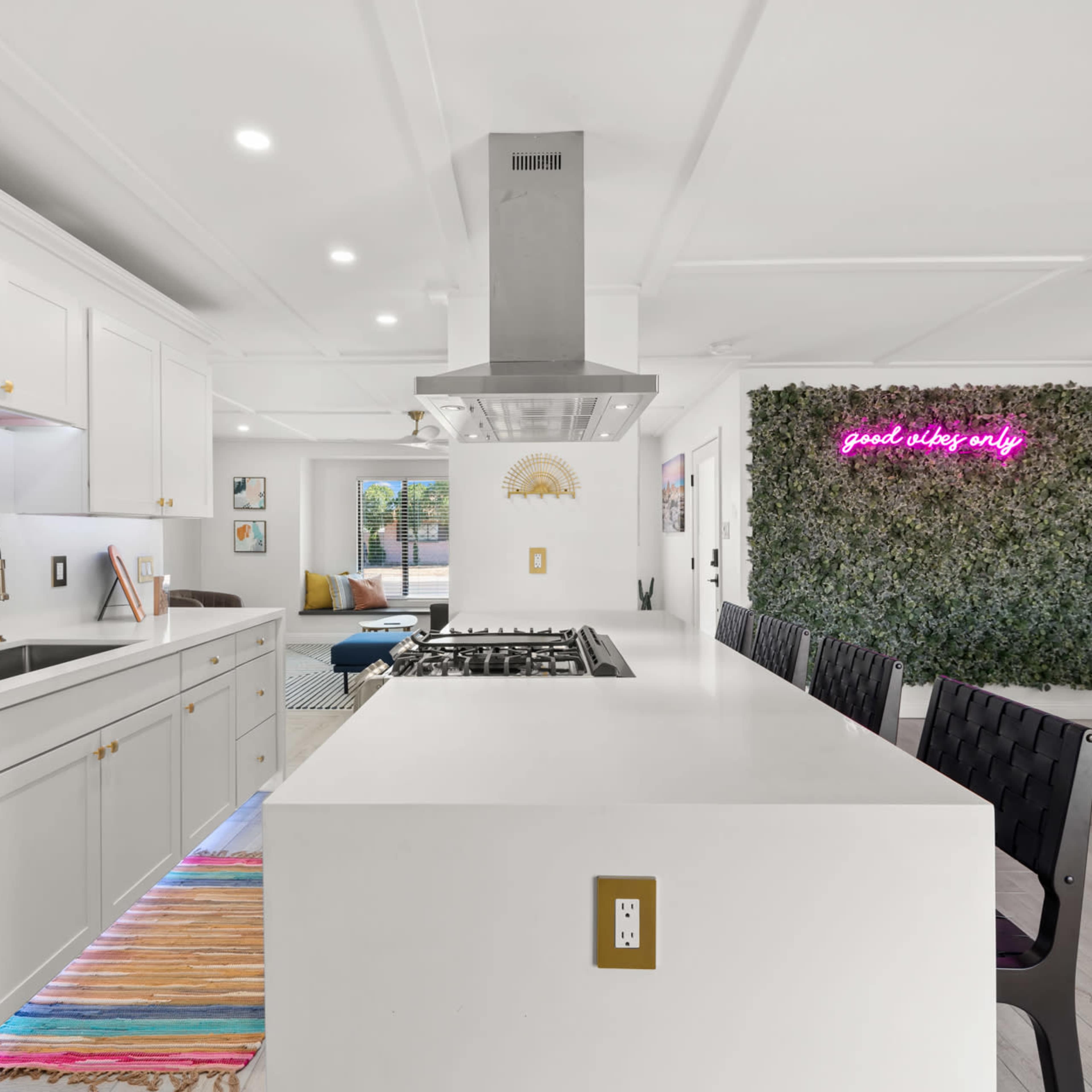 A modern kitchen features white cabinetry, a large island with a gas stove, and a wall adorned with greenery and neon lighting.