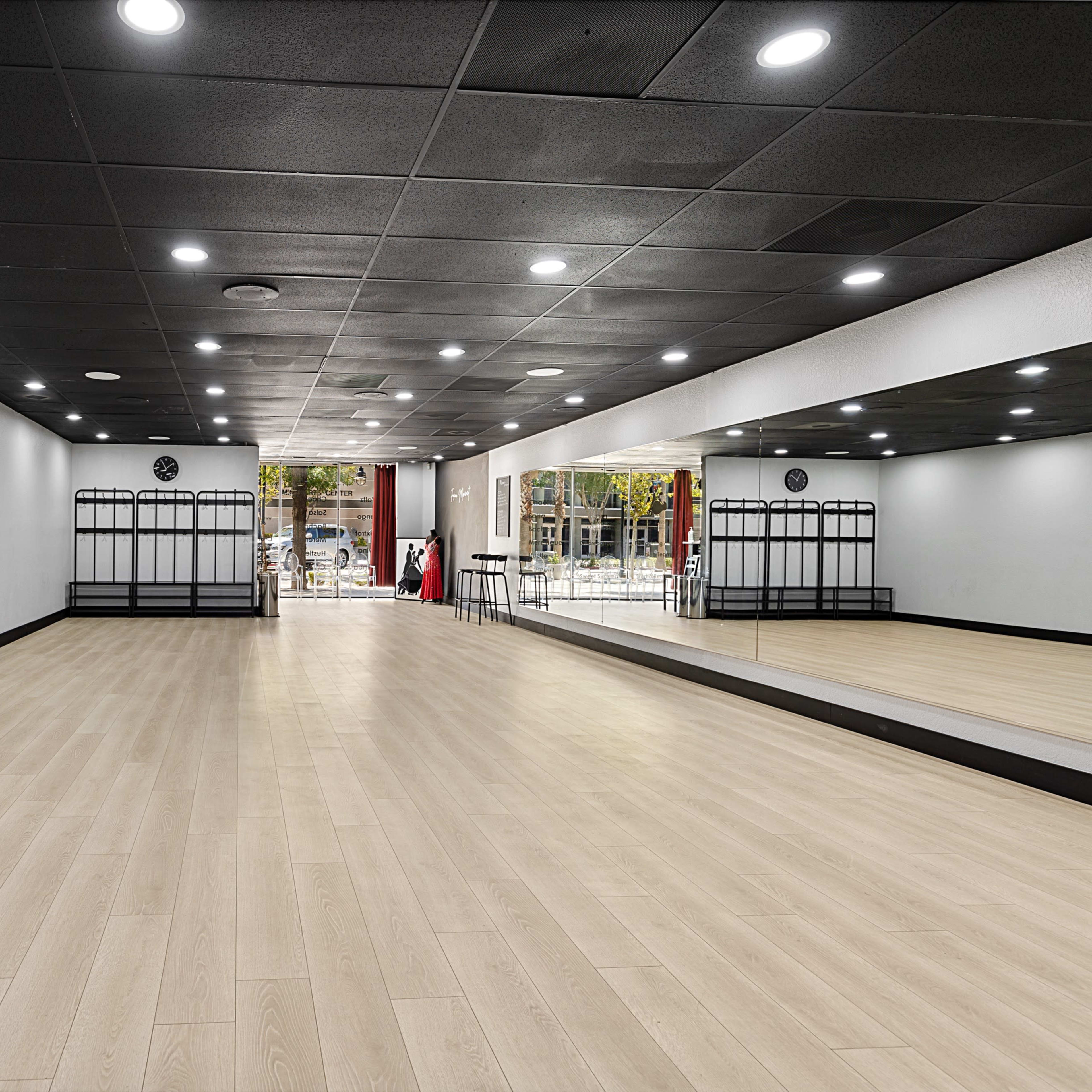 An empty dance studio with wooden flooring, mirrored walls, and a few coat racks along one side.
