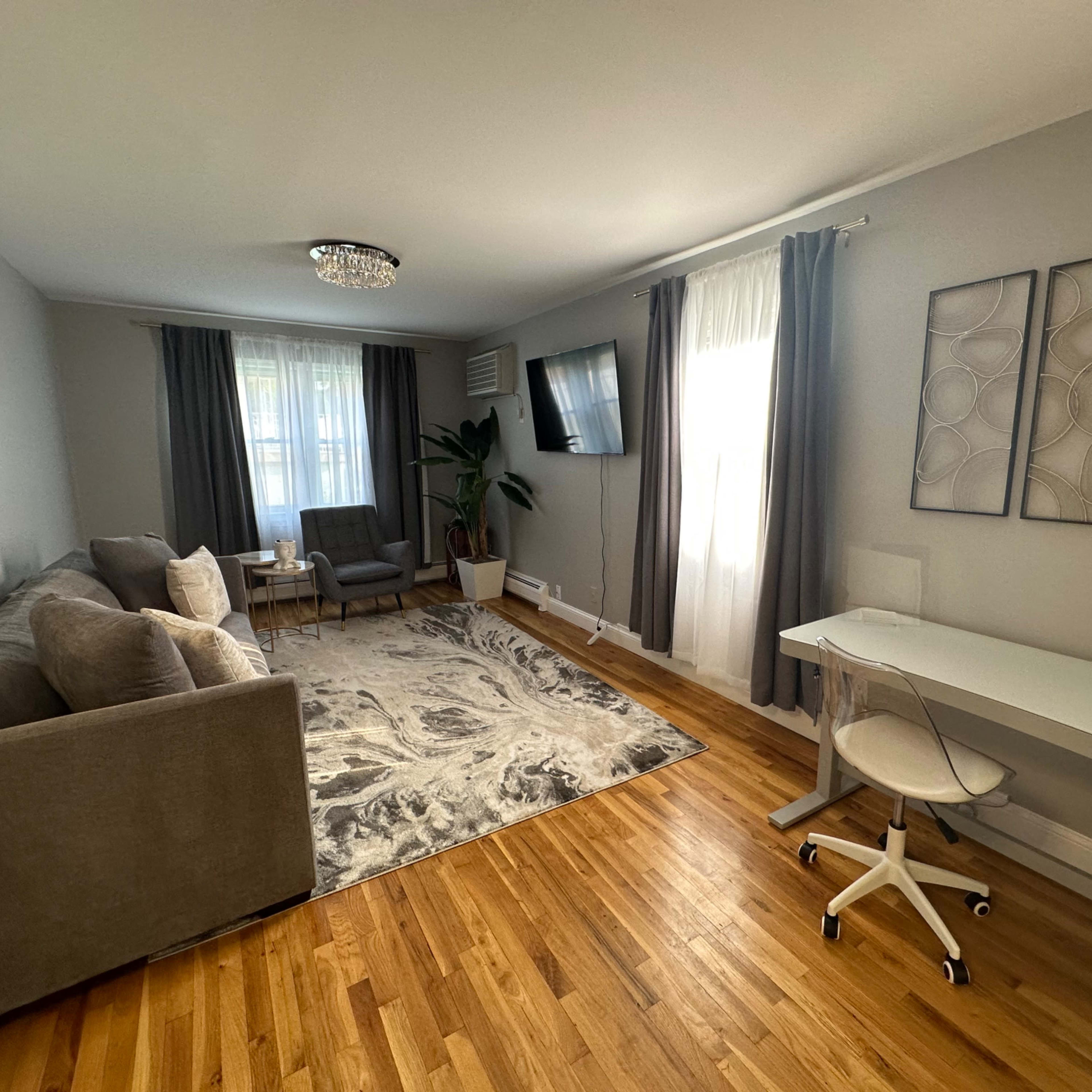 A living room with a gray sofa, a small sitting area, a desk, and a large window with light curtains.