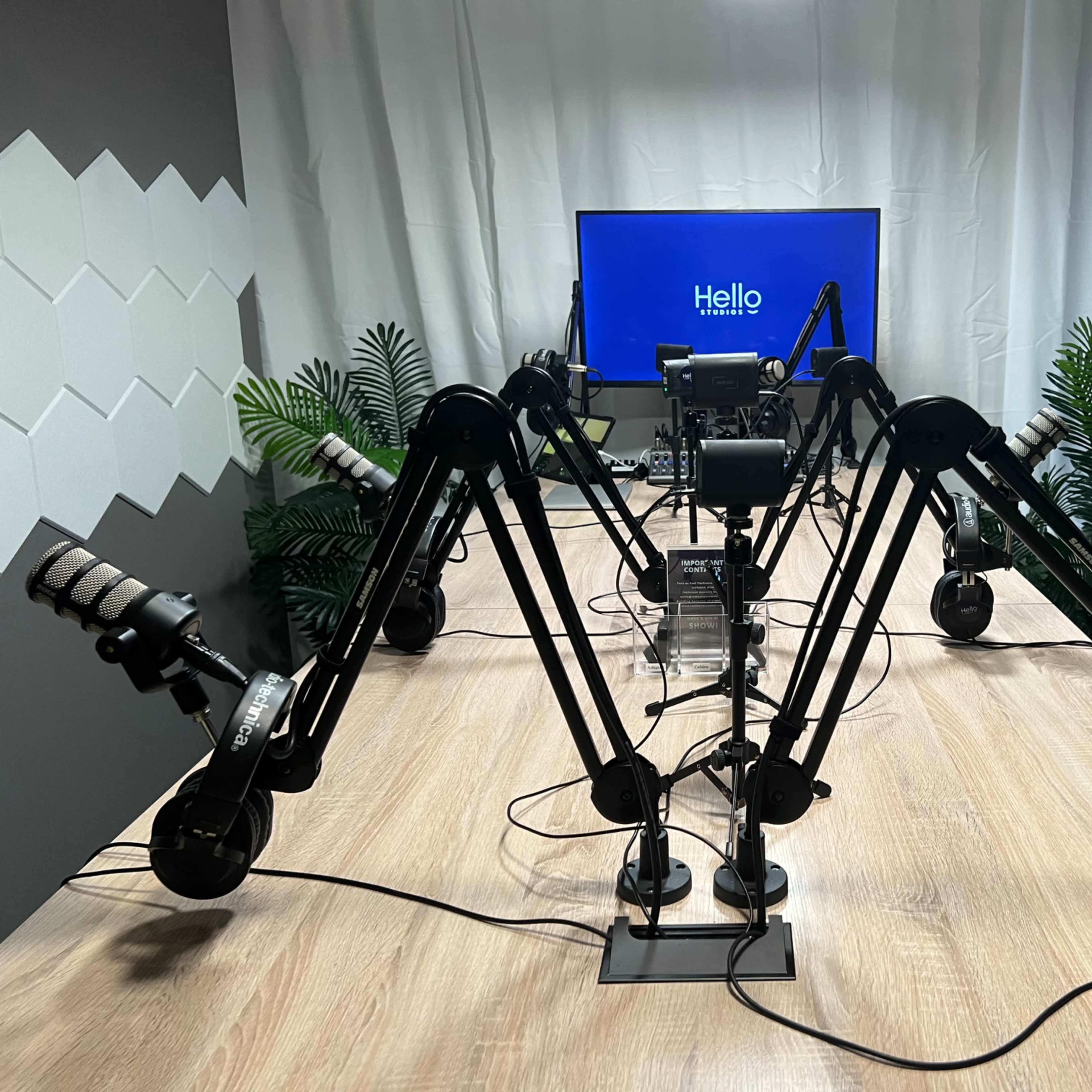 A podcasting setup features several adjustable microphone arms arranged around a central screen with the word "Hello" displayed, against a backdrop of greenery.