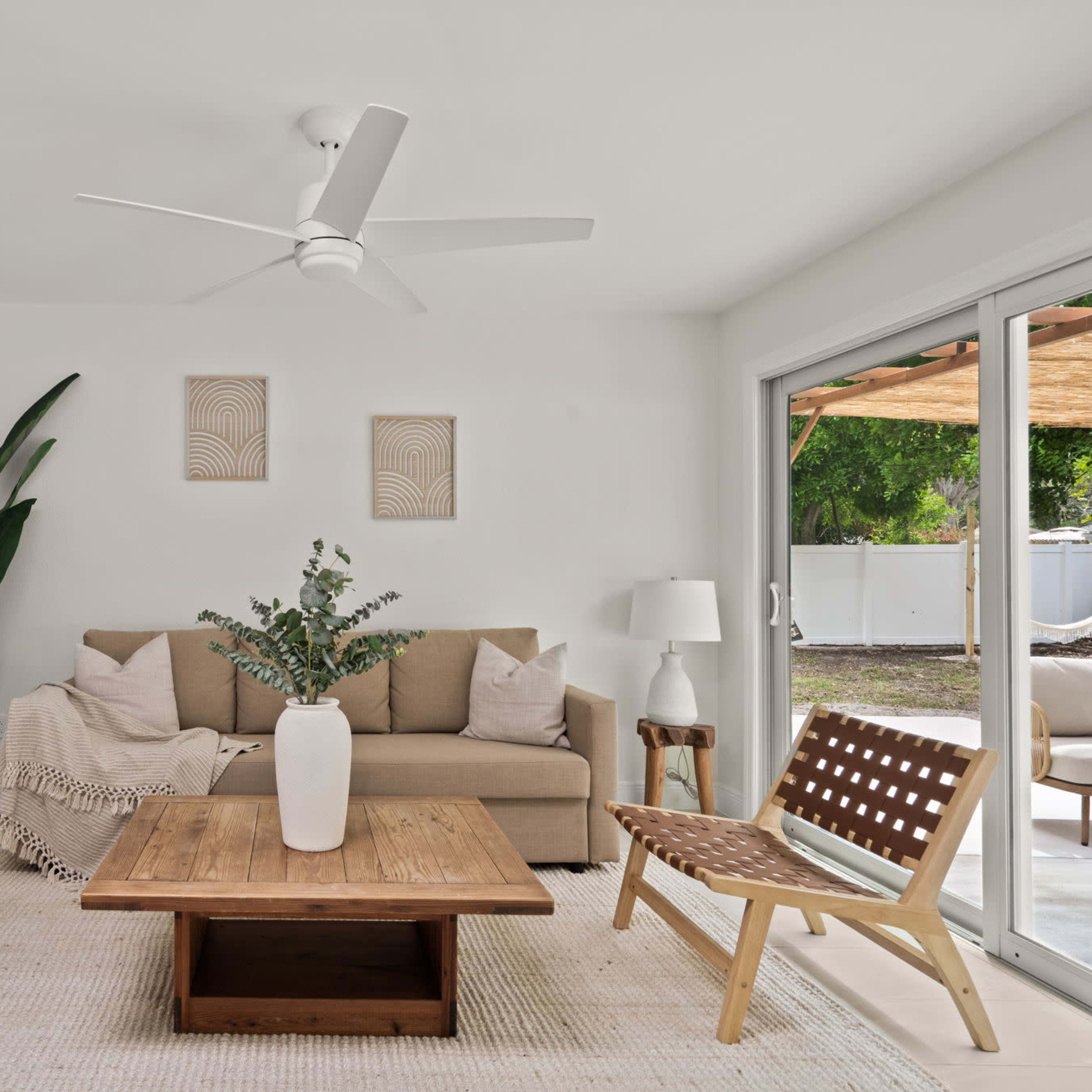 A modern living room features a beige sofa, two wooden chairs with woven seats, a wooden coffee table, and potted plants, illuminated by natural light from sliding glass doors.