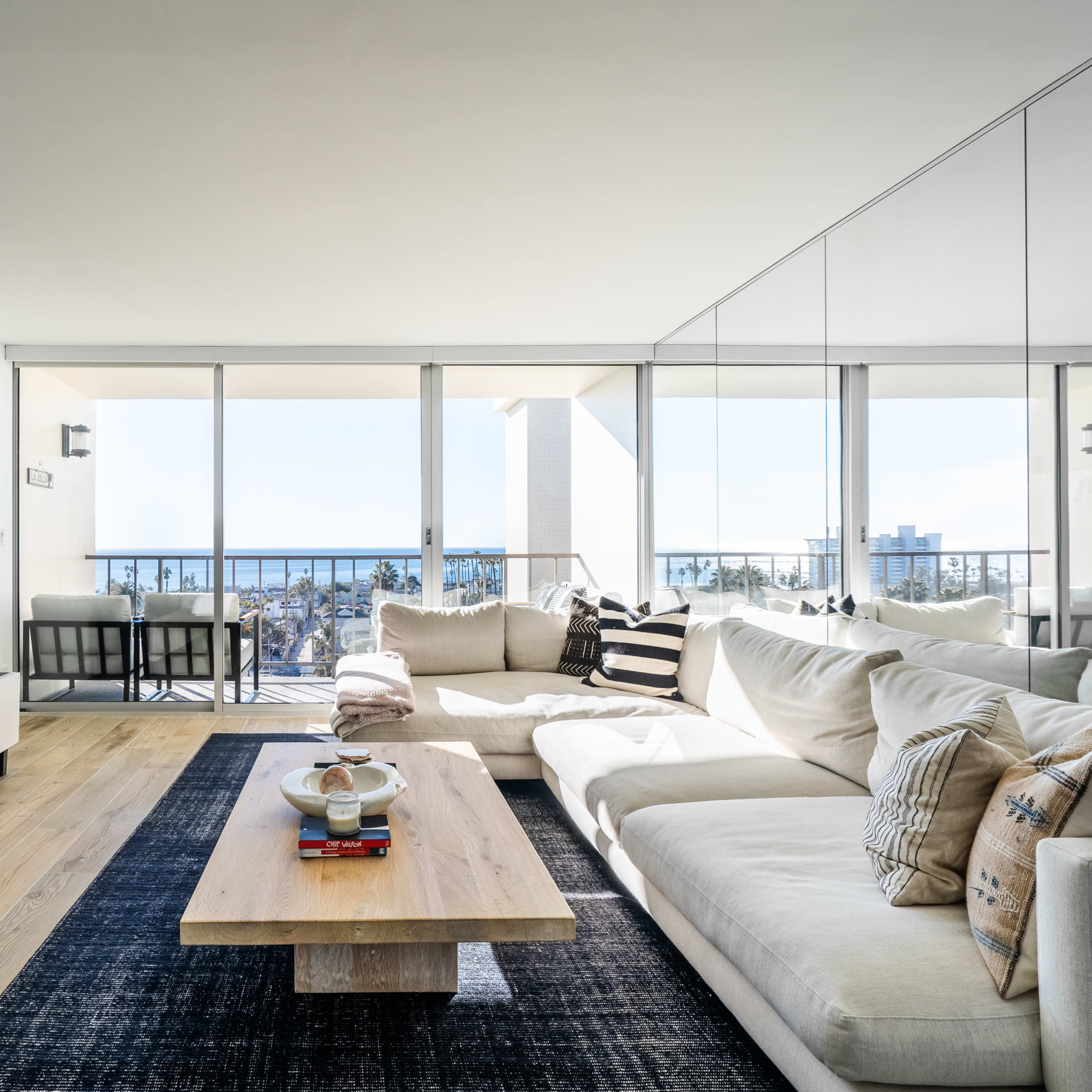 A modern living room features a large sectional sofa, a low wooden coffee table, and a wall-mounted television, with expansive glass doors offering a view of a city skyline.