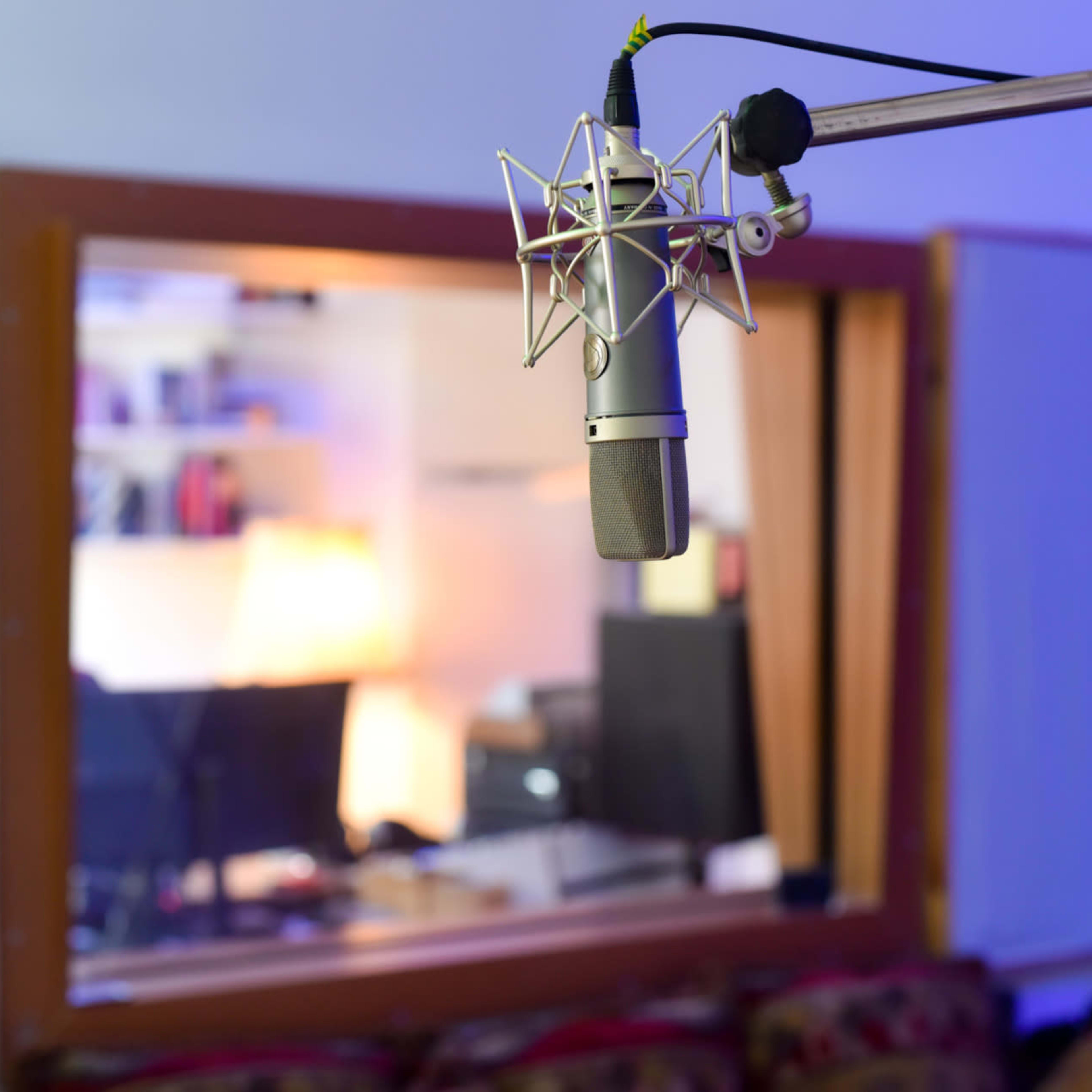 A microphone is positioned in front of a glass window revealing a recording studio with visible monitors and furniture.