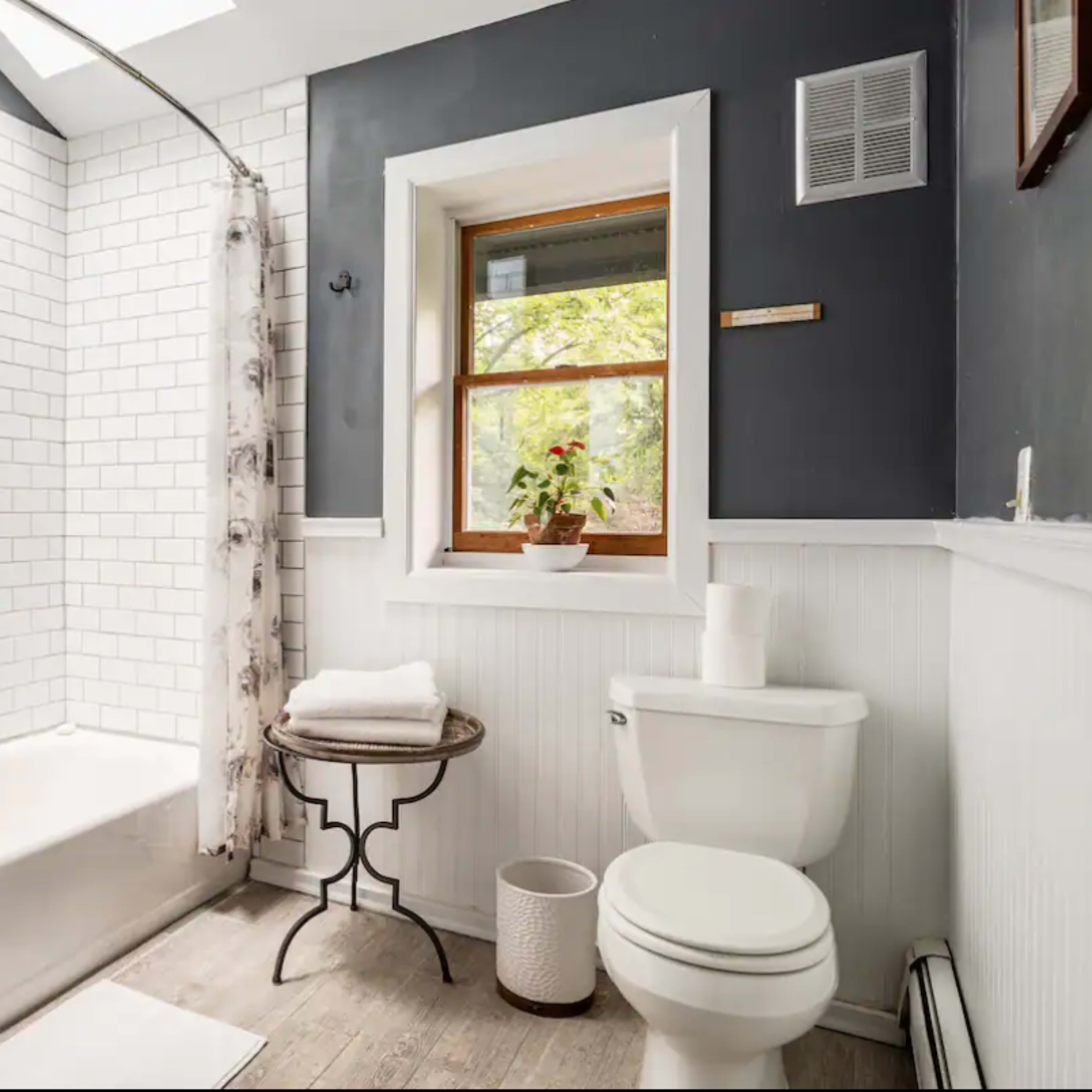 The bathroom features a bathtub with white subway tile, a toilet, a window with a potted plant, and a small table holding neatly stacked towels.
