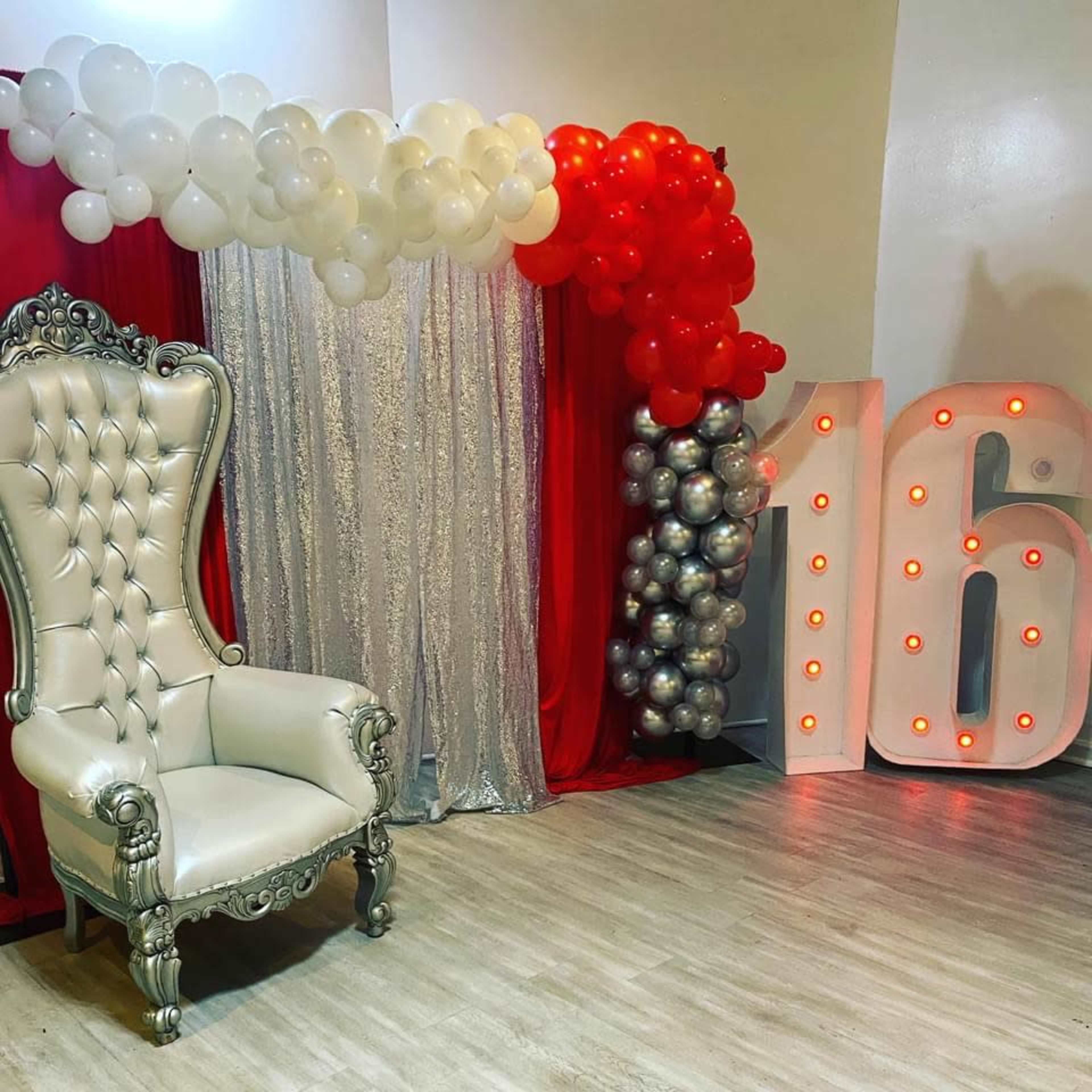 A decorated party area featuring a large silver and red balloon arch, a shimmering silver backdrop, and a plush white throne placed next to a prominent "16" display with lights.