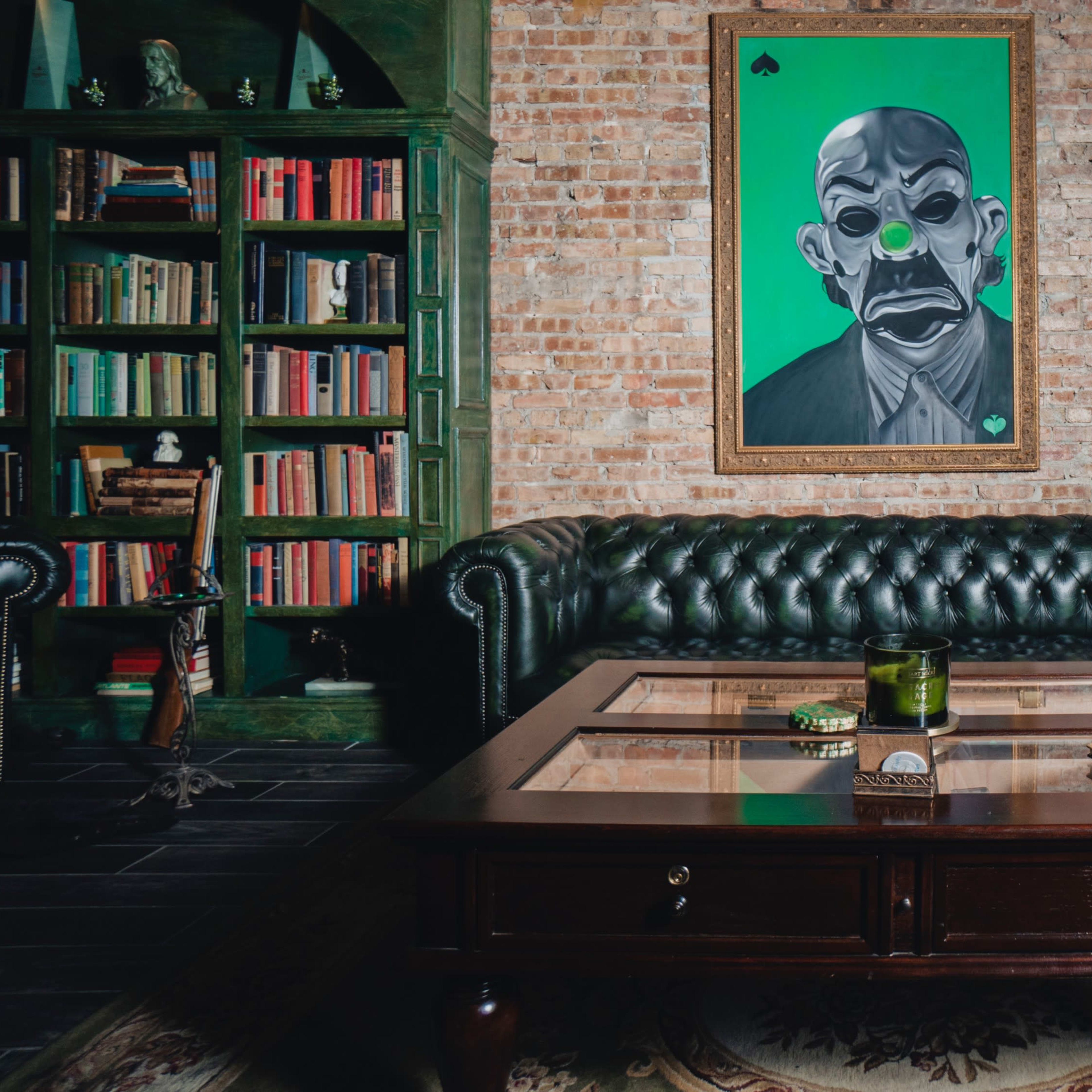 A leather tufted sofa faces a wooden coffee table on a patterned rug, with a colorful portrait on the brick wall above and bookshelves to the left.