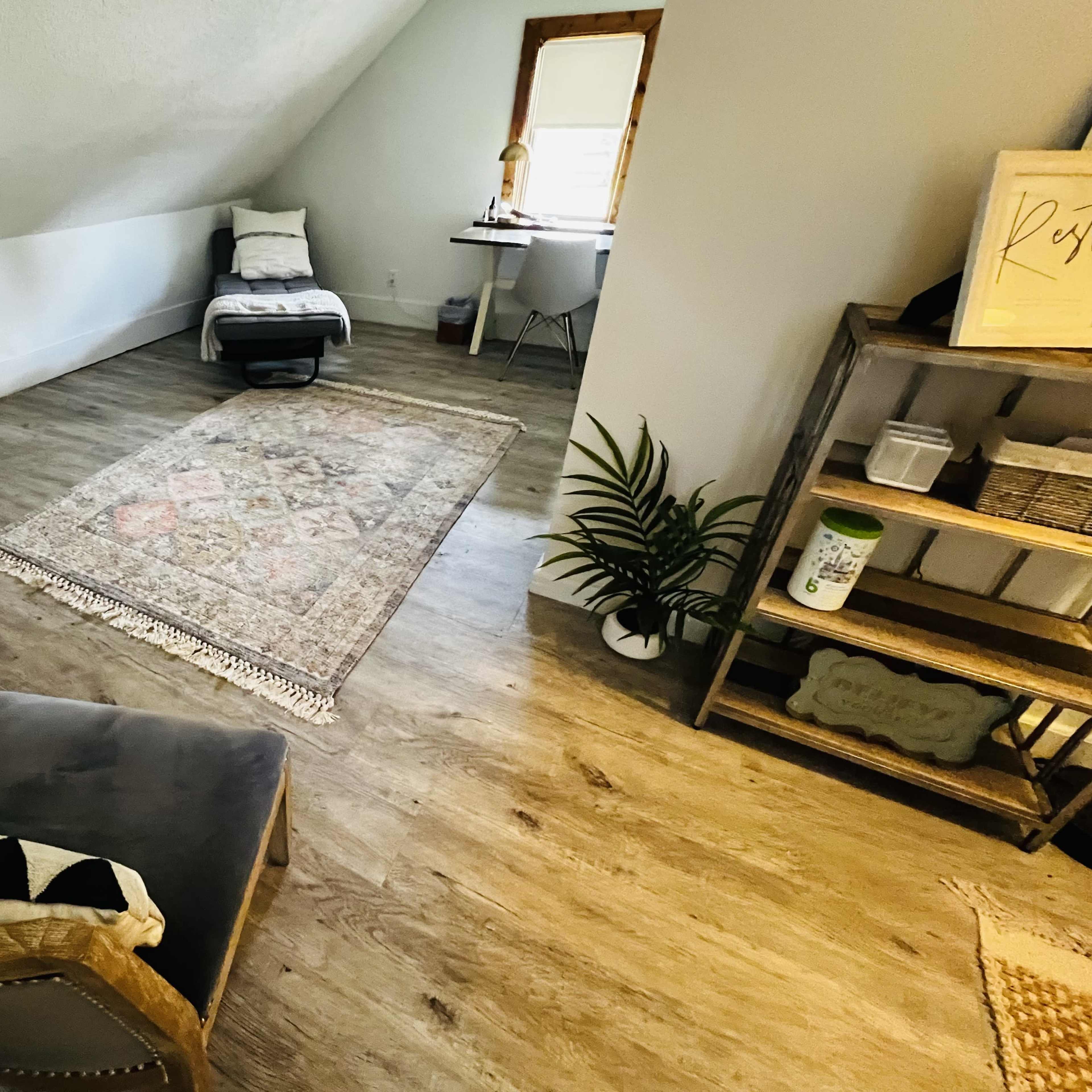 An attic space furnished with a rug, chairs, a small table, and a plant near a window.