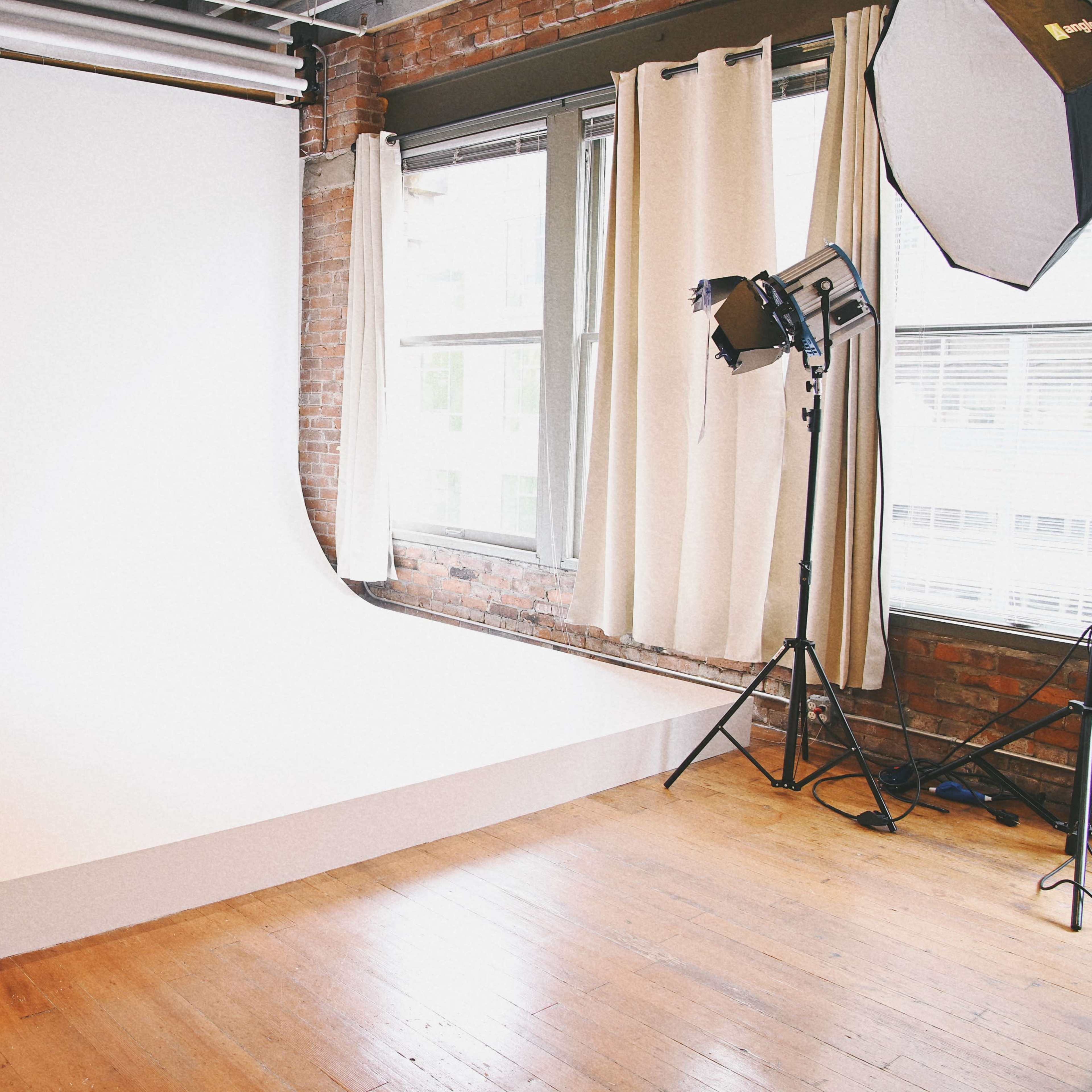 A minimalist photography studio with a seamless white backdrop, wooden flooring, and large windows allowing natural light.